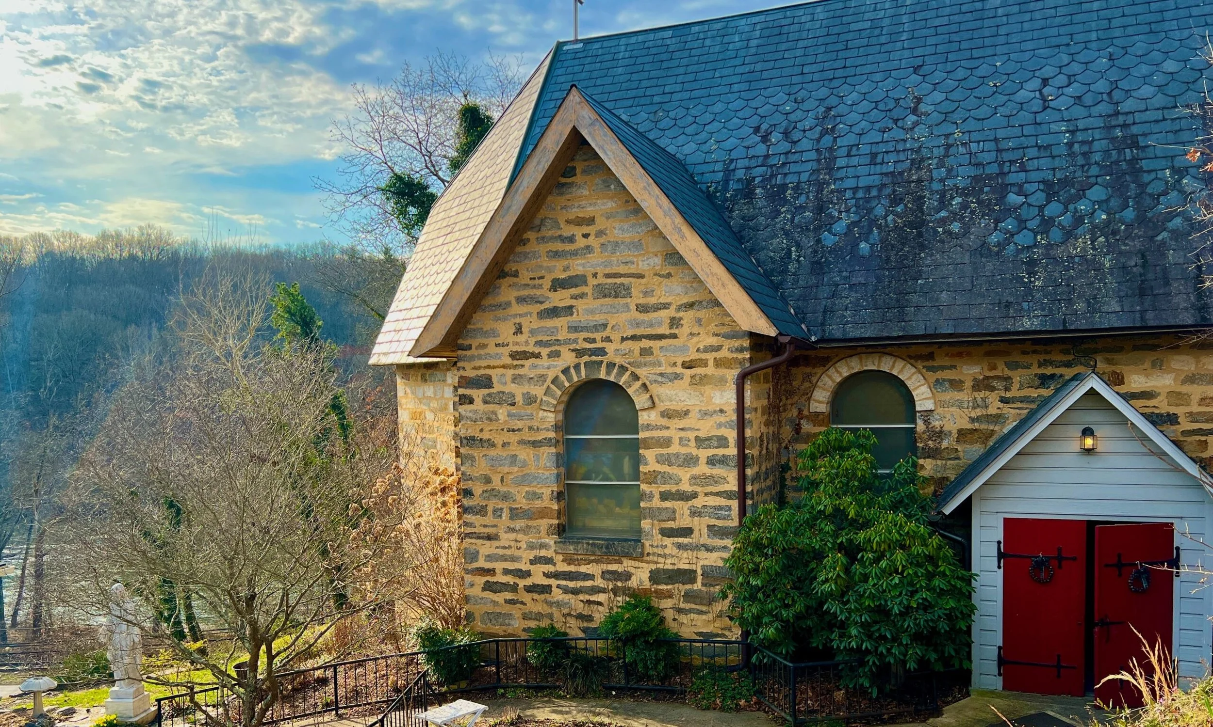 A stone church with arched windows and a steeply sloped roof, next to a small white building with red doors, surrounded by trees and landscaping.