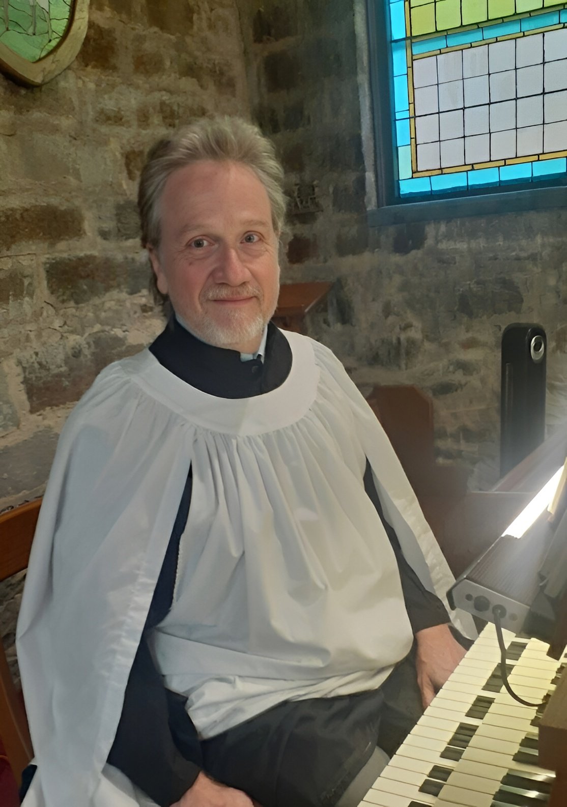 A man dressed as a priest or choir member sitting at a piano inside a stone church with a stained glass window in the background.