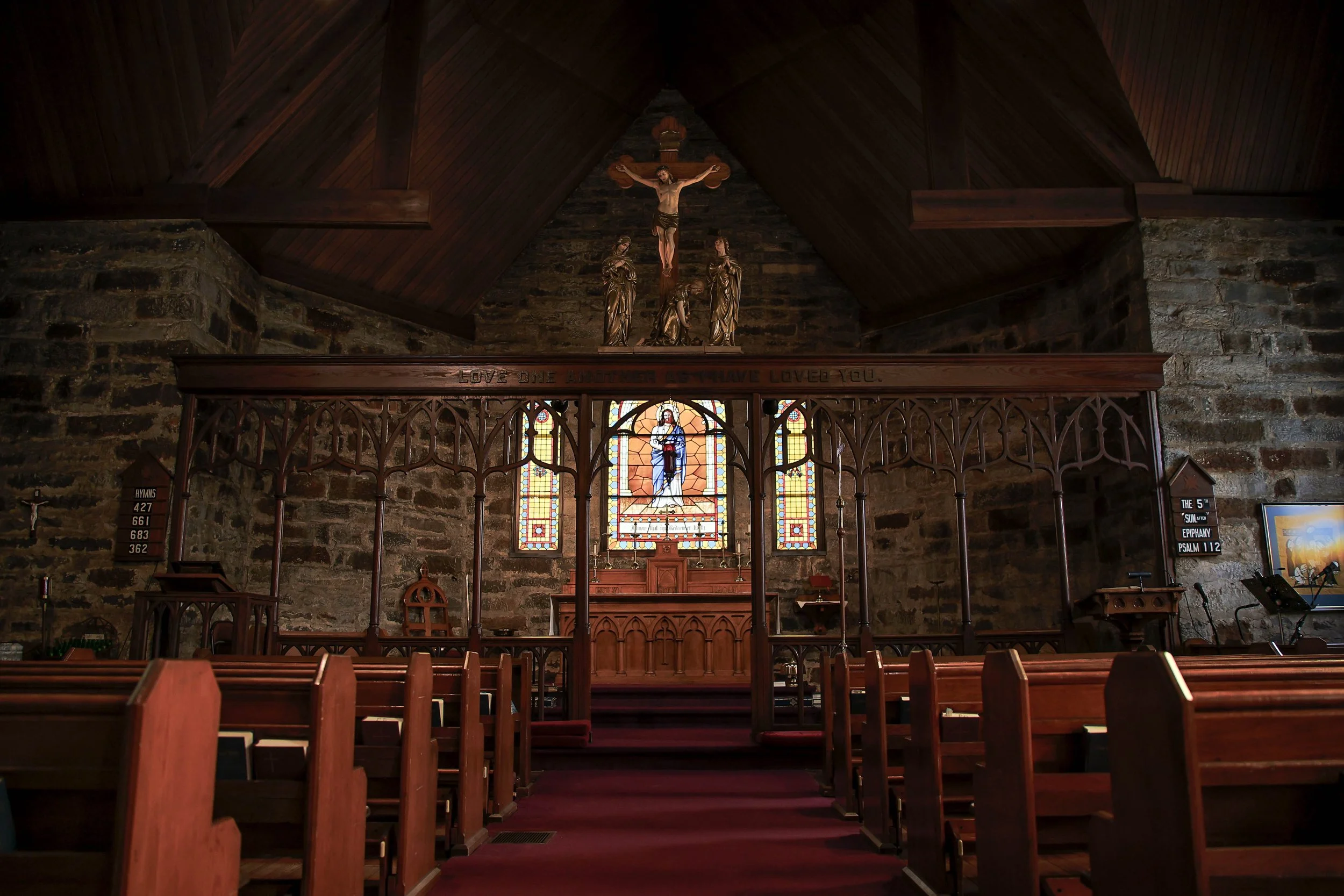 Interior of a church with wooden pews, stained glass windows depicting Jesus, a crucifix, and religious statues above.