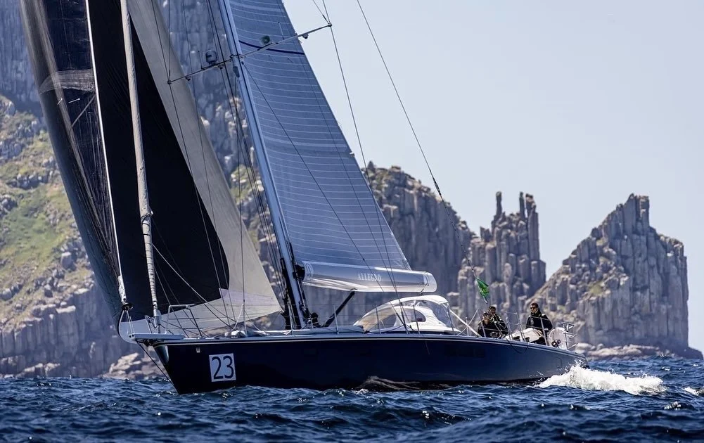 A black sailing yacht with solar panels on the sails, sailing in the ocean near rocky cliffs with people on board.