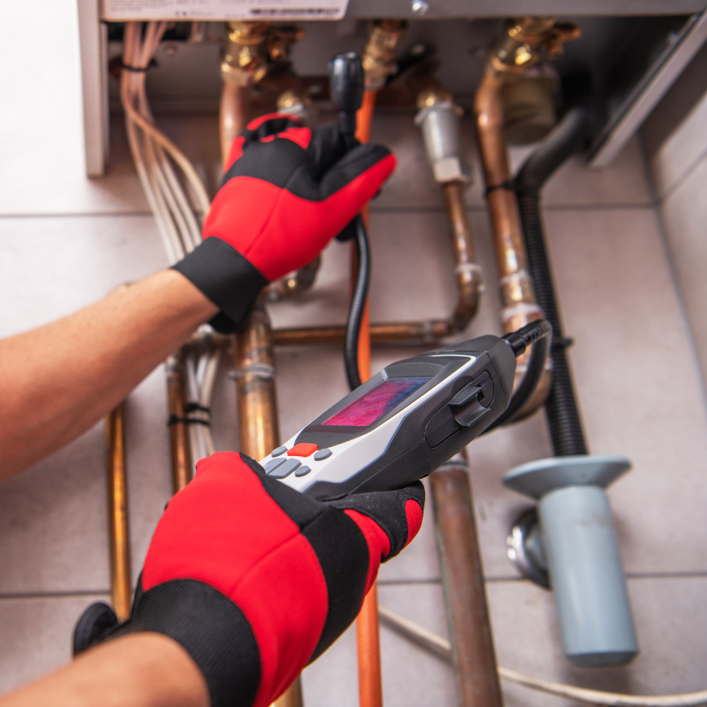 A plumber wearing red and black gloves using a digital multimeter to test copper pipes under a kitchen sink.