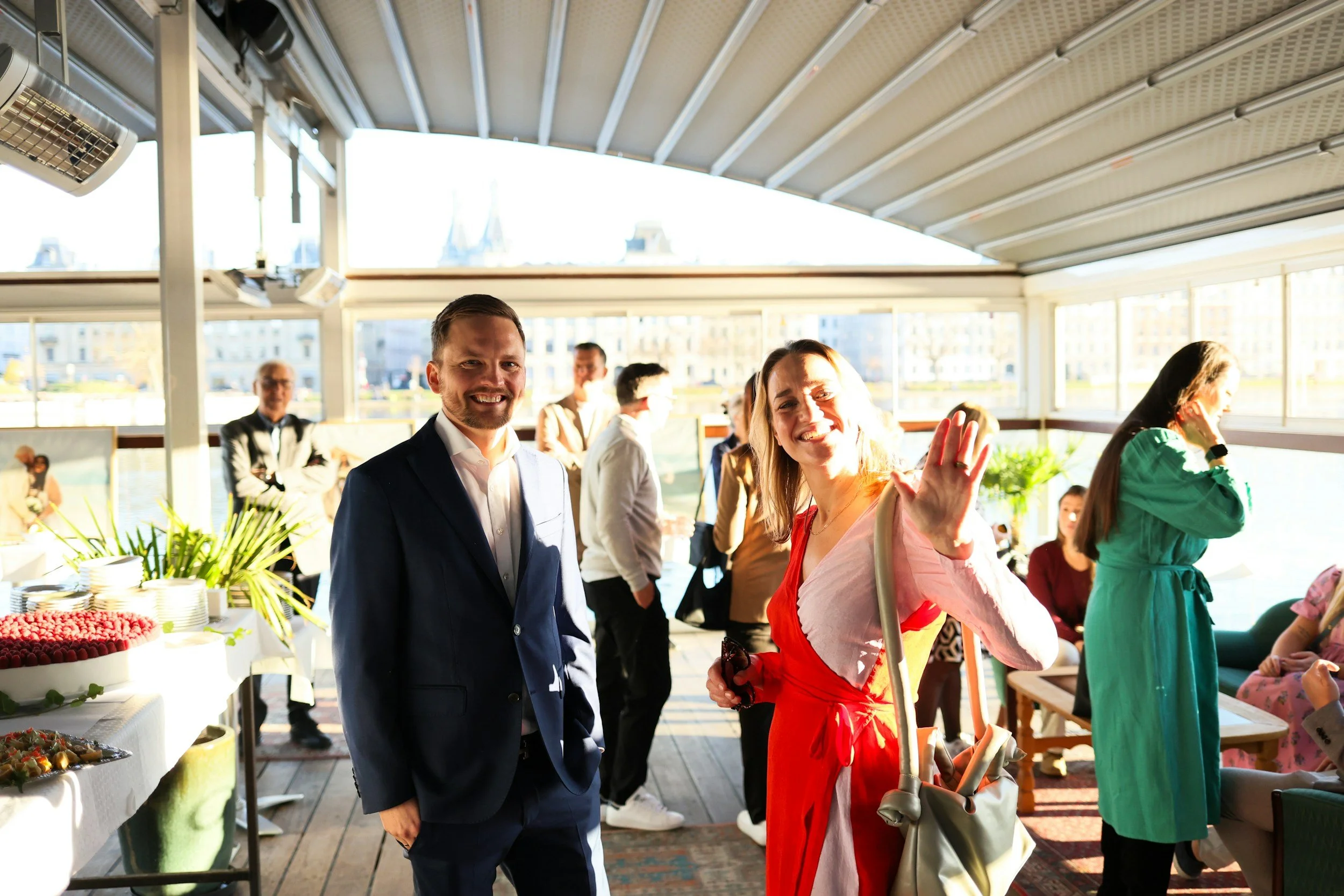 Ihmiset juhlatilaisuudessa, osa taustalla ja buffet-pöytä. Ison ikkunan edessä, aurinko paistaa sisään.