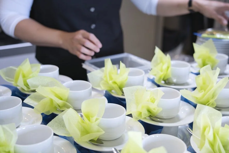 Lautasella on useita pienikokoisia valkoisia kuppeja, joissa on keltaisia paperimaisia koristeita. Taustalla näkyy osa ihmistä, joka valmistaa tai asettelee kuppeja.