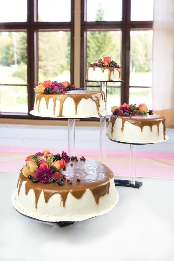 Neljä kakkua, joissa on suklaakerros päällä ja koristeena marjoja ja kukkia, aseteltuna pöydälle pitkän säpäleisen ikkunan edessä.
