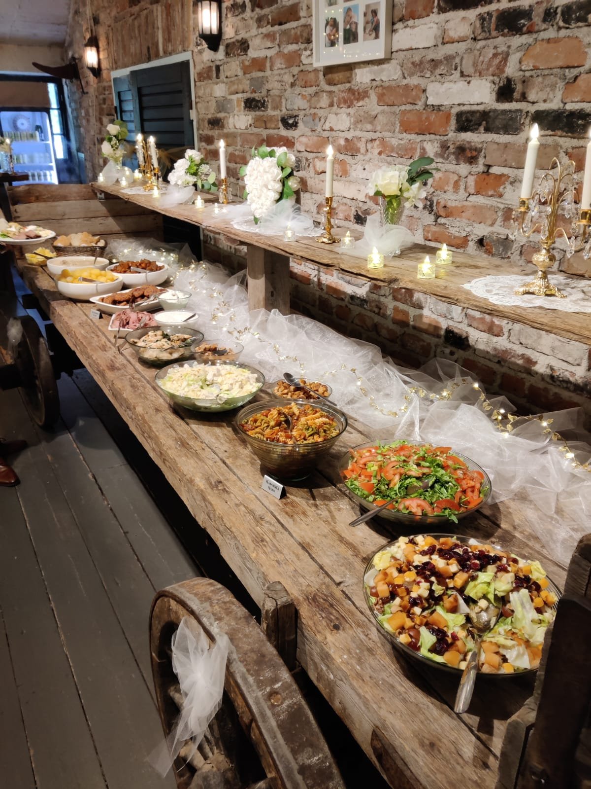 Lautasen ruokapöytä, jossa on erilaisia salaatteja ja kylmiä ruokia, koristeltu kynttilöillä ja kukkivilla kukkavazoilla. Taustalla on tiilirakenteinen seinä ja hylly, jossa on valokuvakehyksiä.