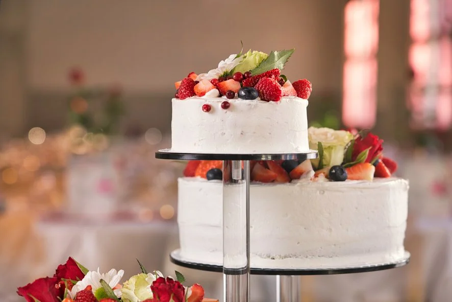 Hääkakku, koristeltu erilaisilla tuoreilla marjoilla ja hedelmillä, kahdella tasolla, aseteltu keskelle juhlasalia.