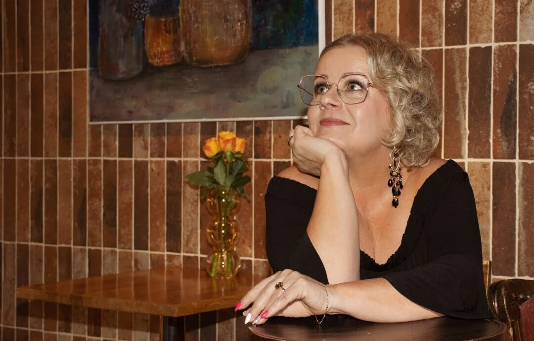 Nainen istuu pöydän ääressä, katsoo ylös ja hymyilee, taustalla kukkakuvioinen seinä ja pöydällä lasimaljallinen keltainen-oransseja kukkia.