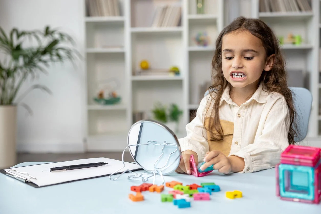 A young girl with curly brown hair, wearing a cream-colored shirt, sitting at a table, playing with colorful letter tiles and looking into a small, round mirror, working on speech sounds during speech therapy for kids in Houston, TX.