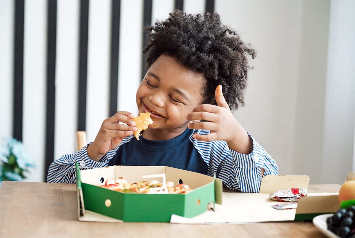 A young boy with curly hair working on trying new foods during feeding therapy for kids in Houston, TX, eating a slice of pizza while sitting at a table with a box of pizza in front of him.