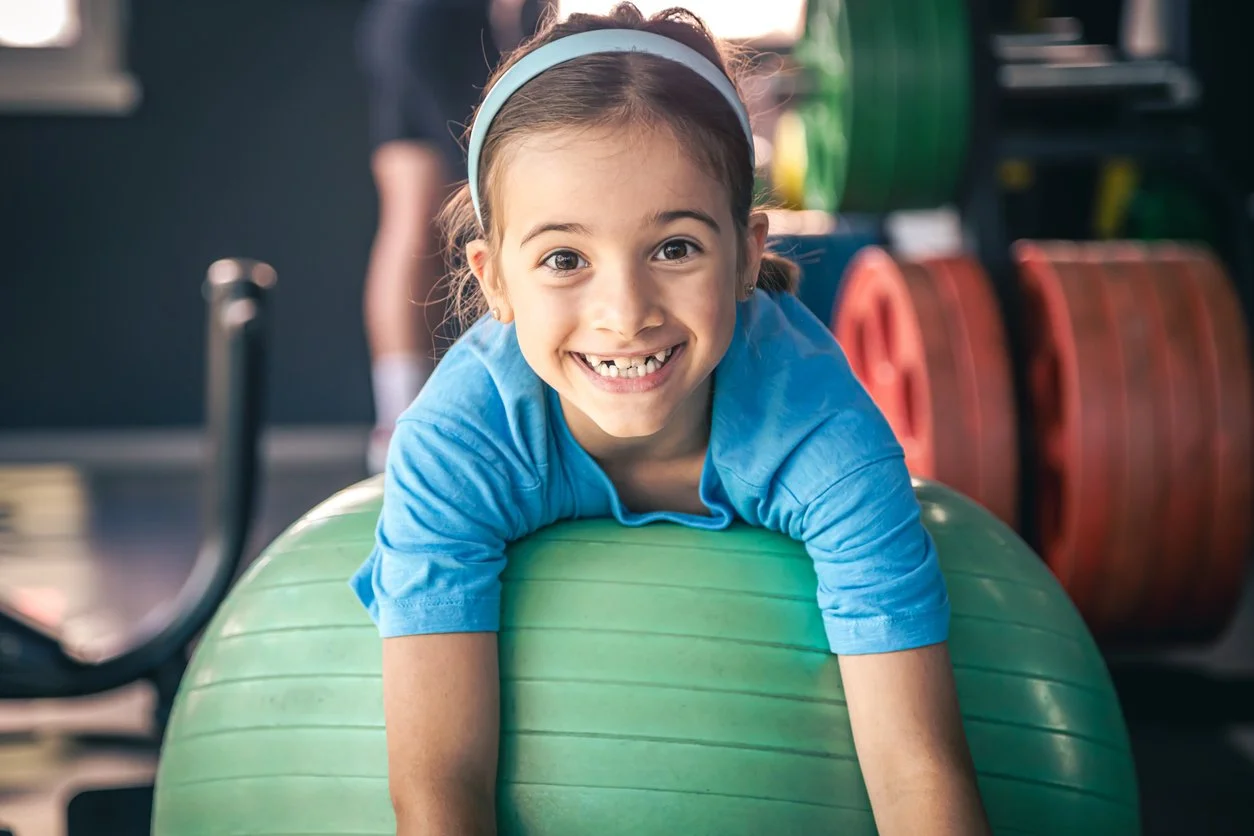 A young girl with a big smile, wearing a blue shirt and a headband, lying on a green exercise ball in a gym during OT for children in Houston, TX.