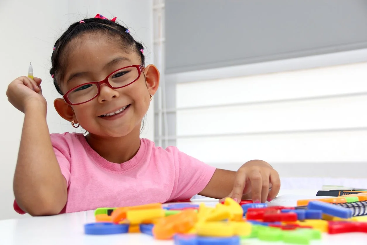 Young girl with glasses smiling and playing with colorful plastic counting coins at a table, working on vocabulary skills and following directions during speech therapy for children in Houston, TX.