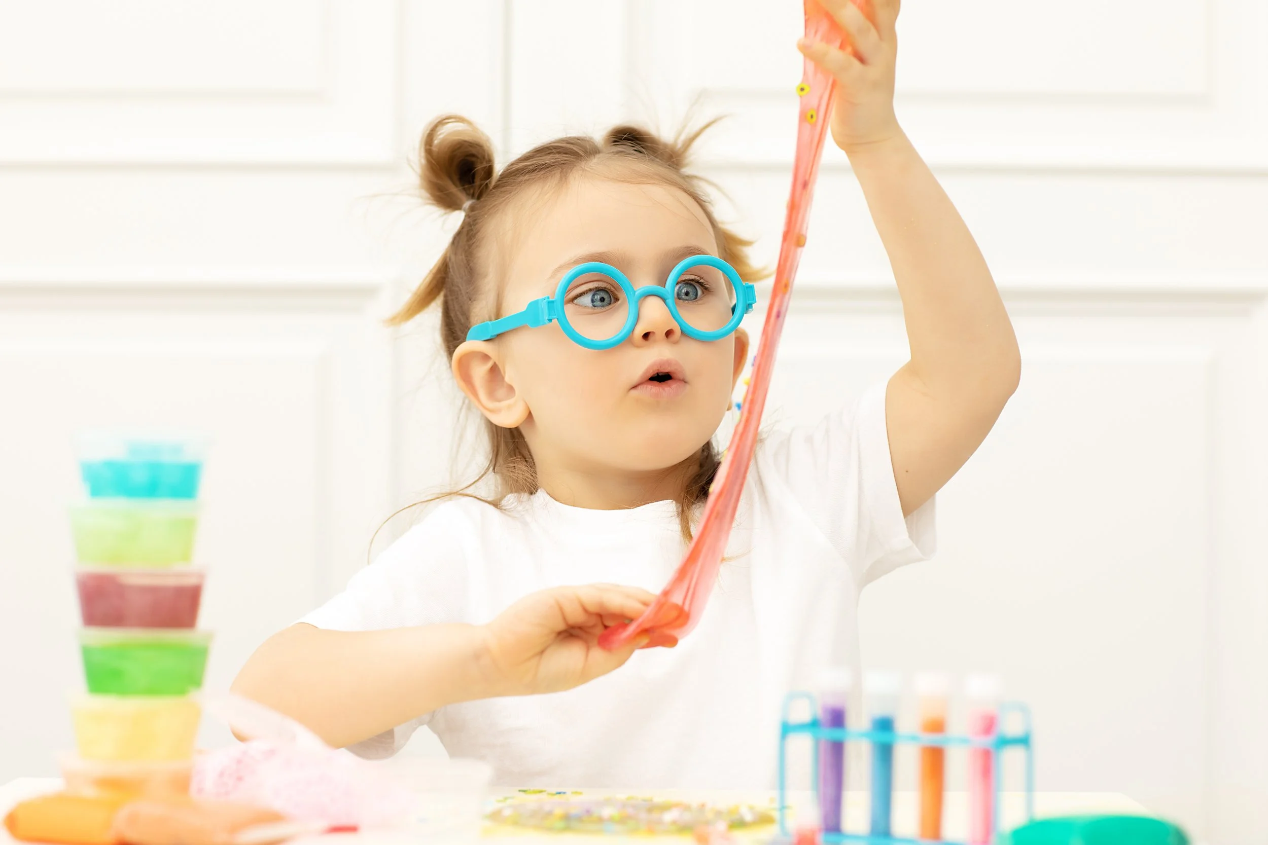 Young girl wearing large blue glasses holding a pink and orange slime, with colorful slime containers and testing tubes on the table, working on sensory and motor skills during occupational therapy for kids in Houston, TX.