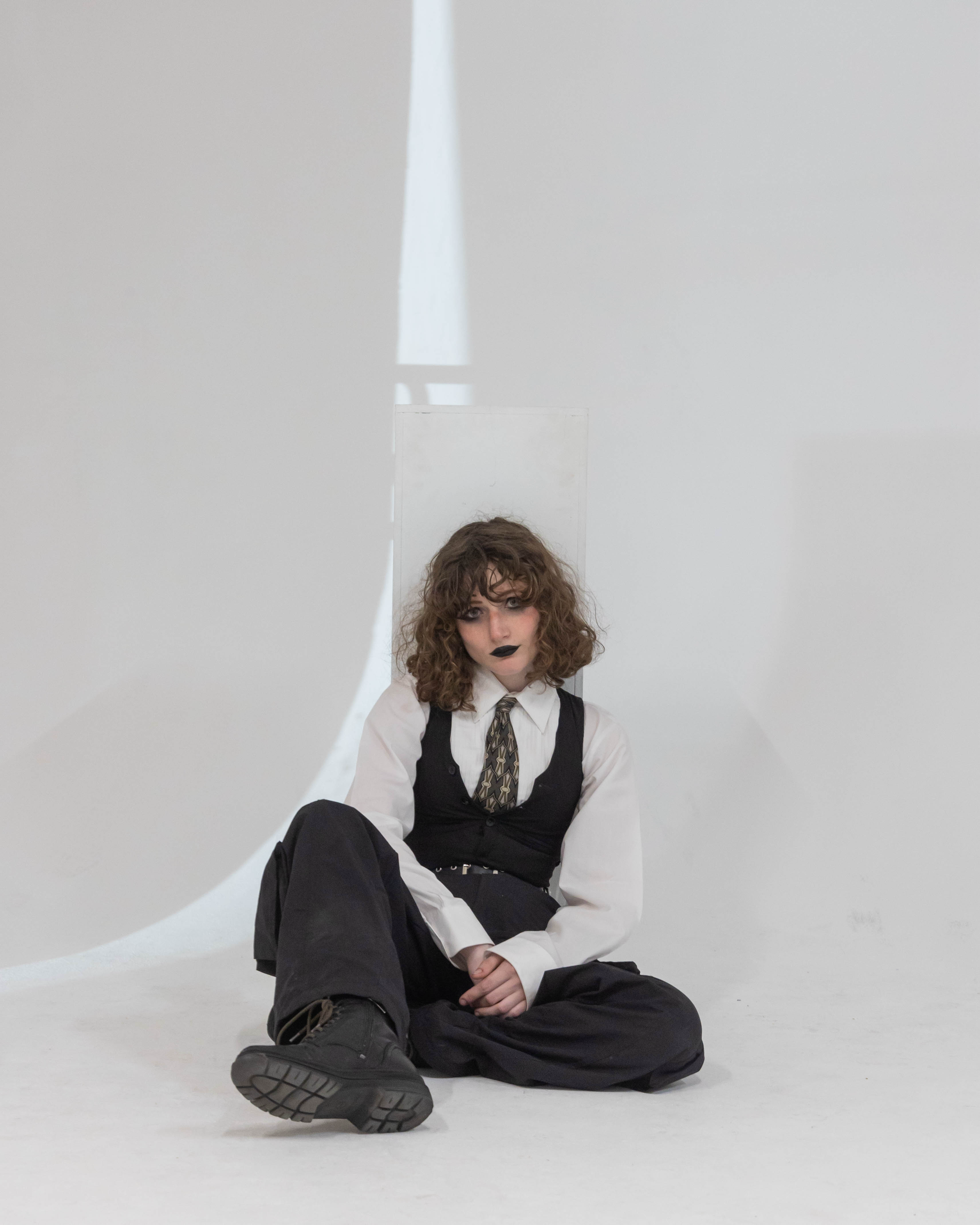 Person with curly hair, dark lipstick, white shirt, black vest, patterned tie, black pants, and boots sitting on the floor against a plain background with a large white curved shape.