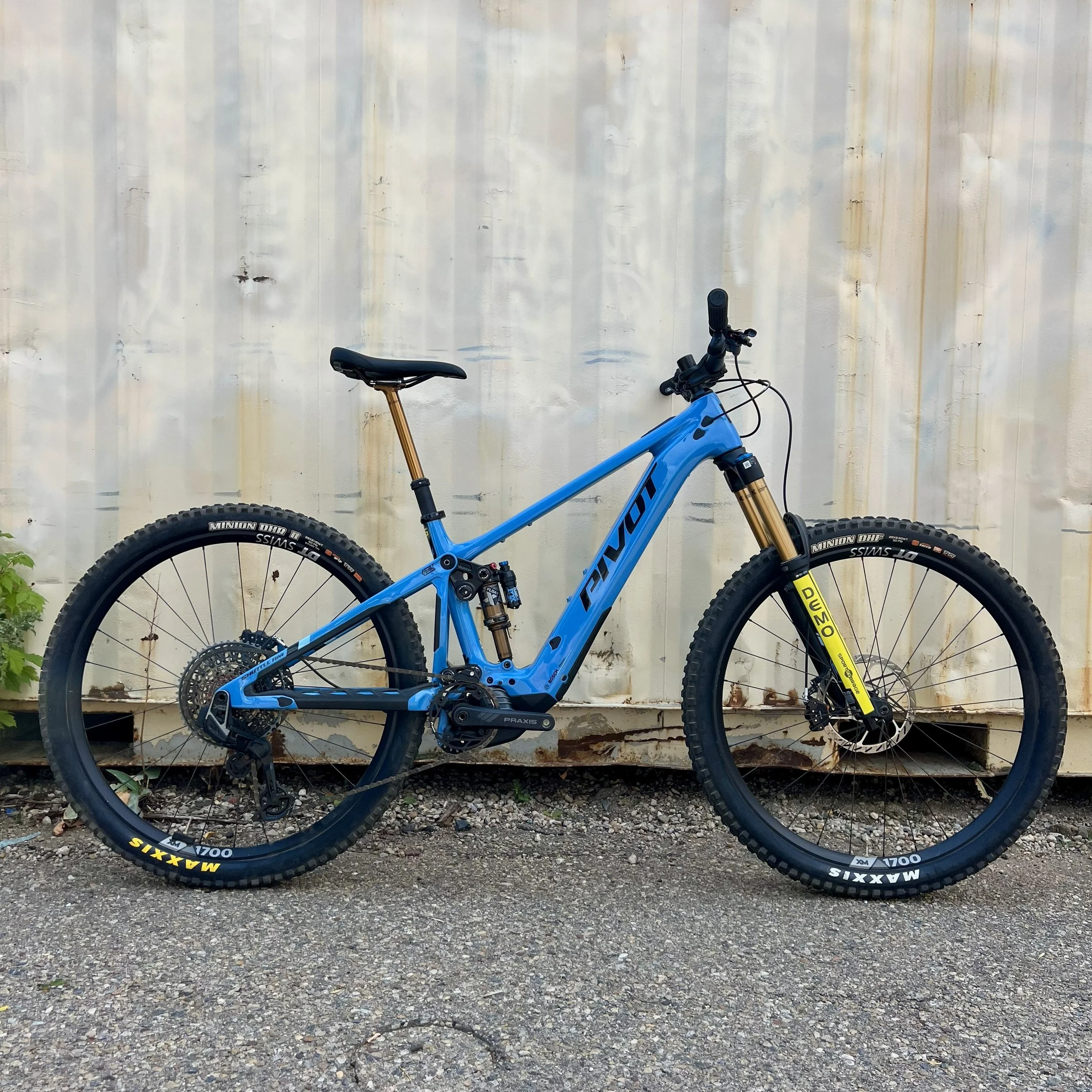 Blue Trek mountain bike with gold suspension forks and Maxxis tires, parked against a weathered, off-white wall.