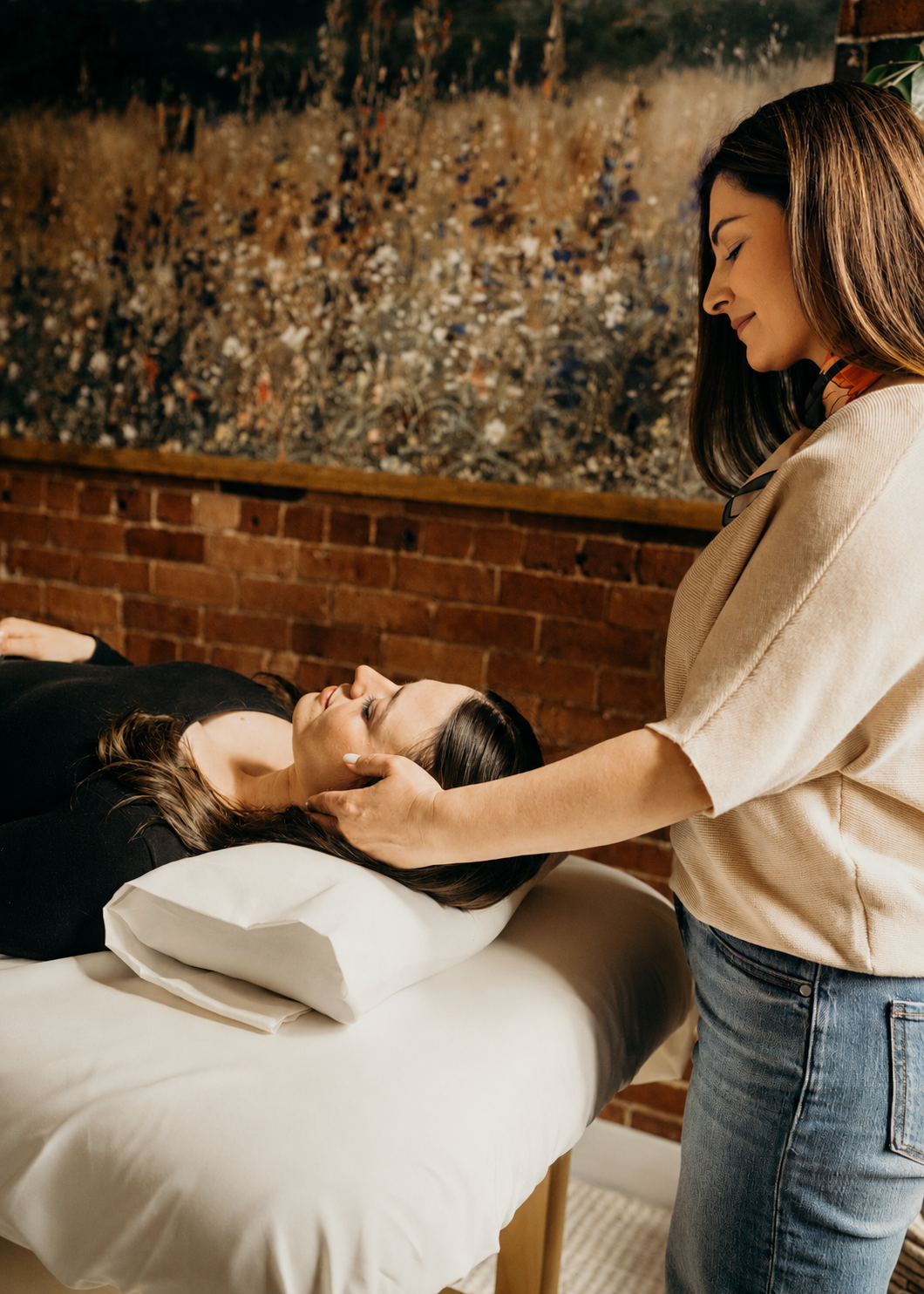 Woman receiving a Reiki energy healing at a wellness center with brick wall and floral art in the background. The practitioner is using energy healing techniques and psychic intuition to facilitate the healing.