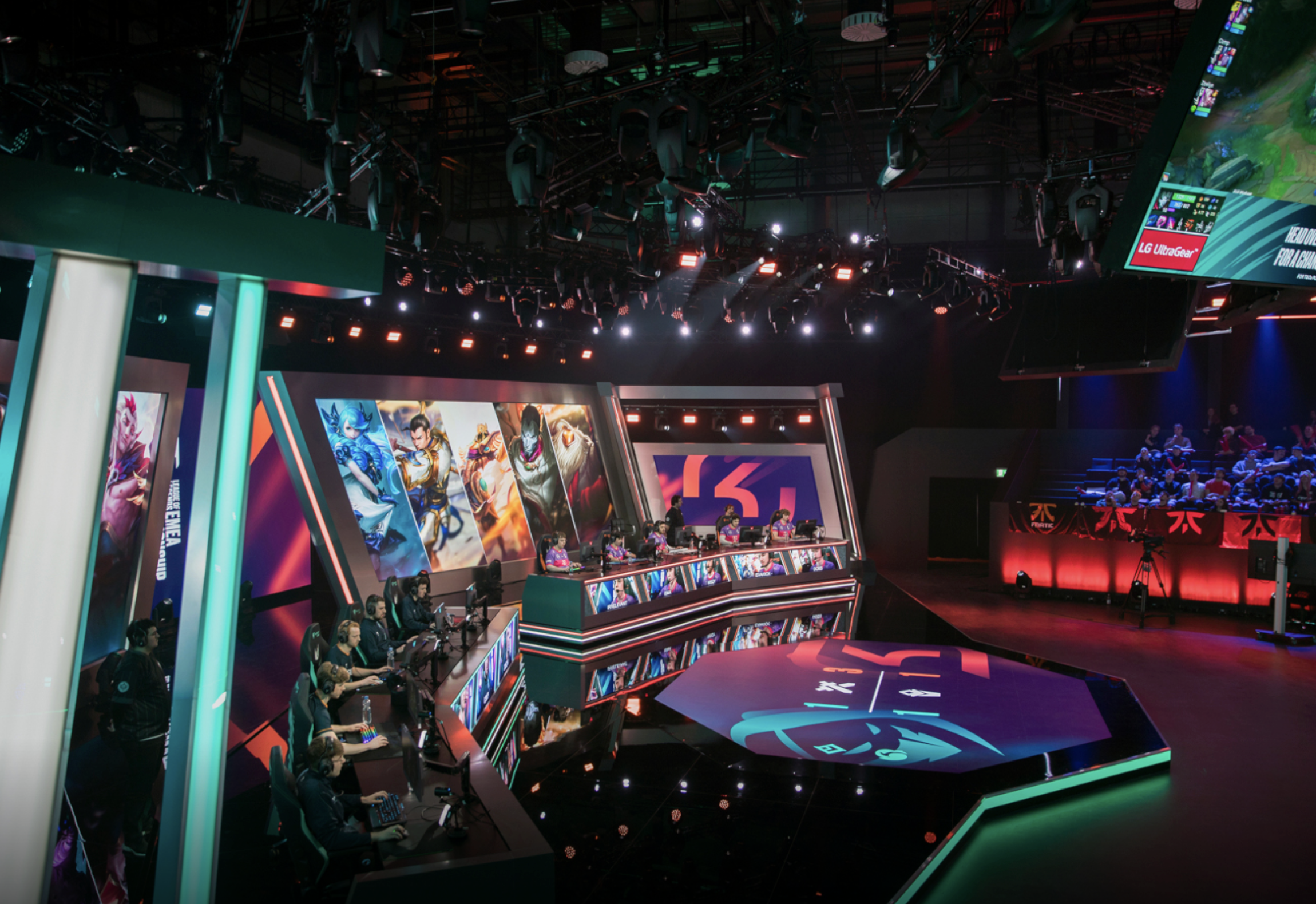 An eSports gaming event with players seated at desks, large screens displaying game images, and an audience watching in the background, illuminated by colorful stage lighting.