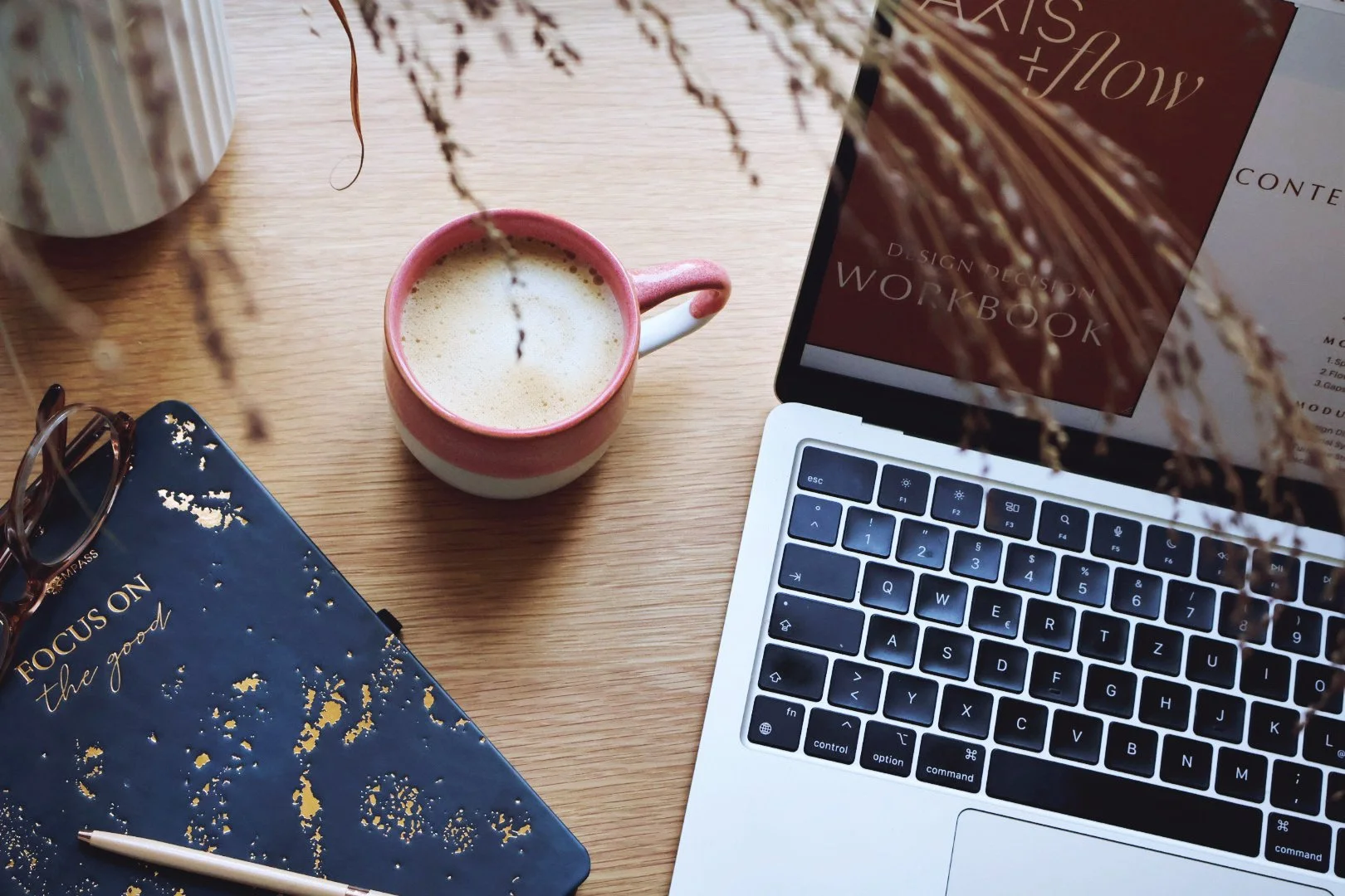 A laptop, a pink and white coffee mug with a frothy drink, a pair of glasses, a blue notebook with gold speckles, a white pen, and a potted plant on a wooden desk.