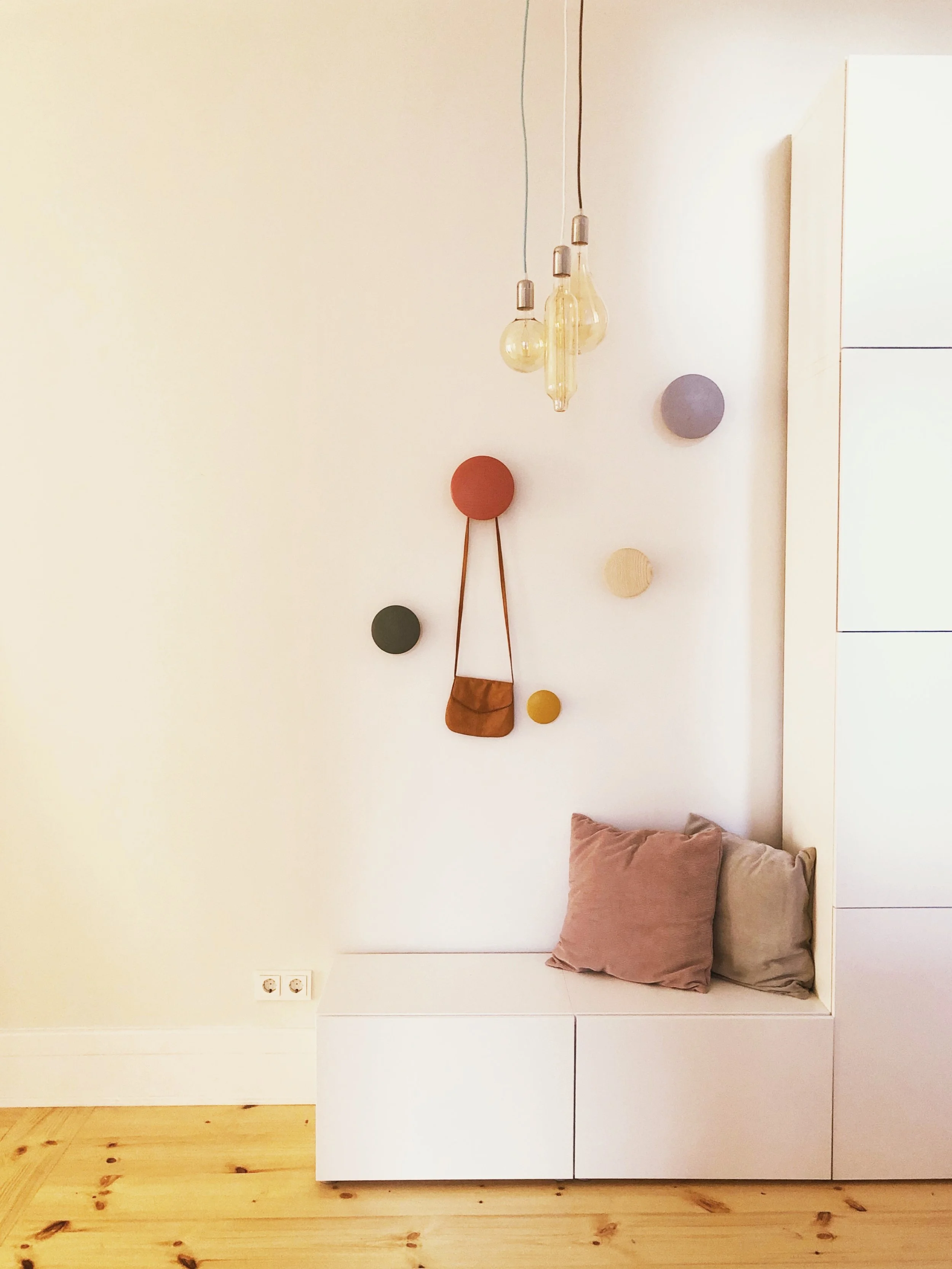 Minimalist entryway with a white bench, two pillows, decorative wall plates, hanging pendant lights, and a small brown bag on the wall.
