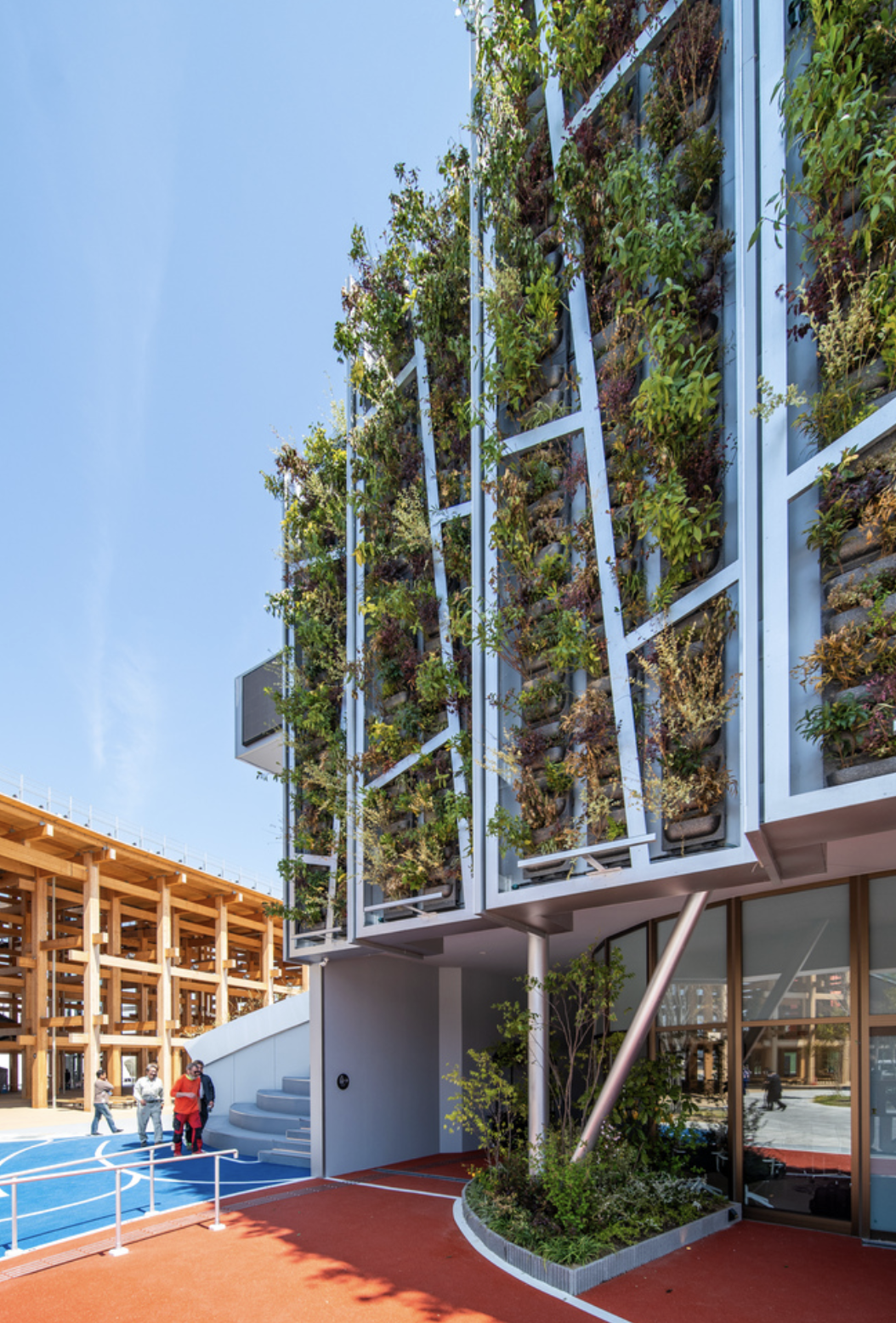 Modern building with vertical garden, people walking on colorful outdoor space, wooden structure in background, clear blue sky.