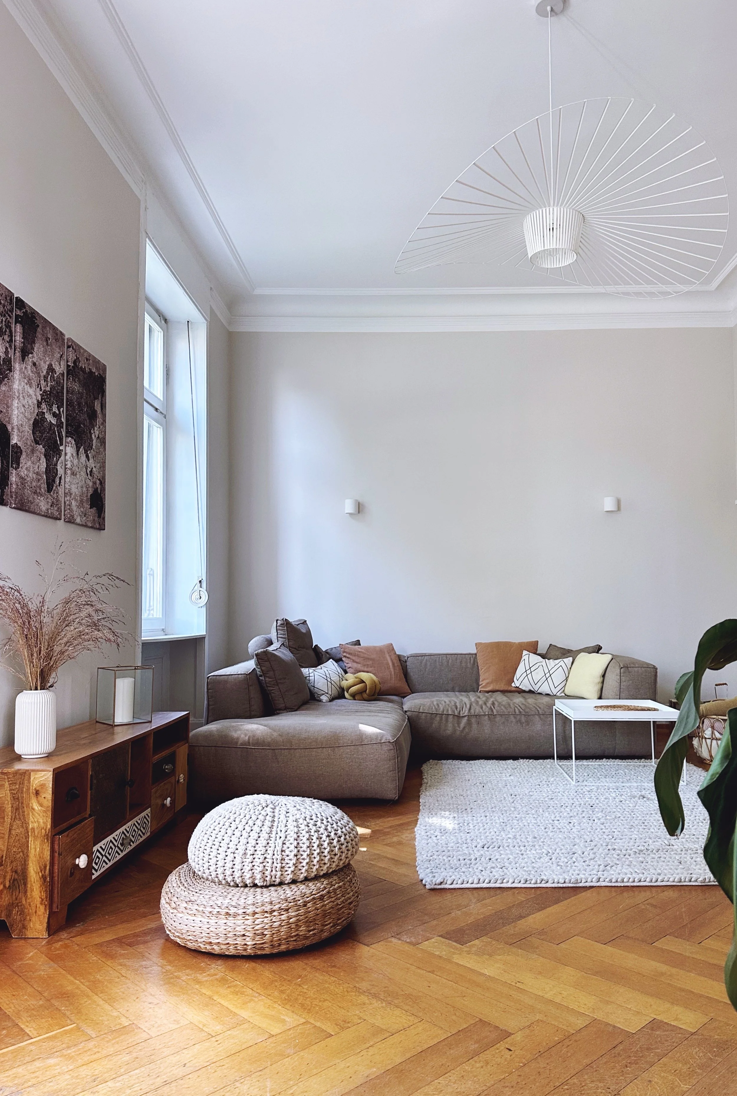 A cozy living room with a sectional sofa, decorative pillows, a white textured rug, a woven pouf, a wooden sideboard with decorative items, a window with sheer curtains, and modern wall art, with a unique ceiling light fixture.