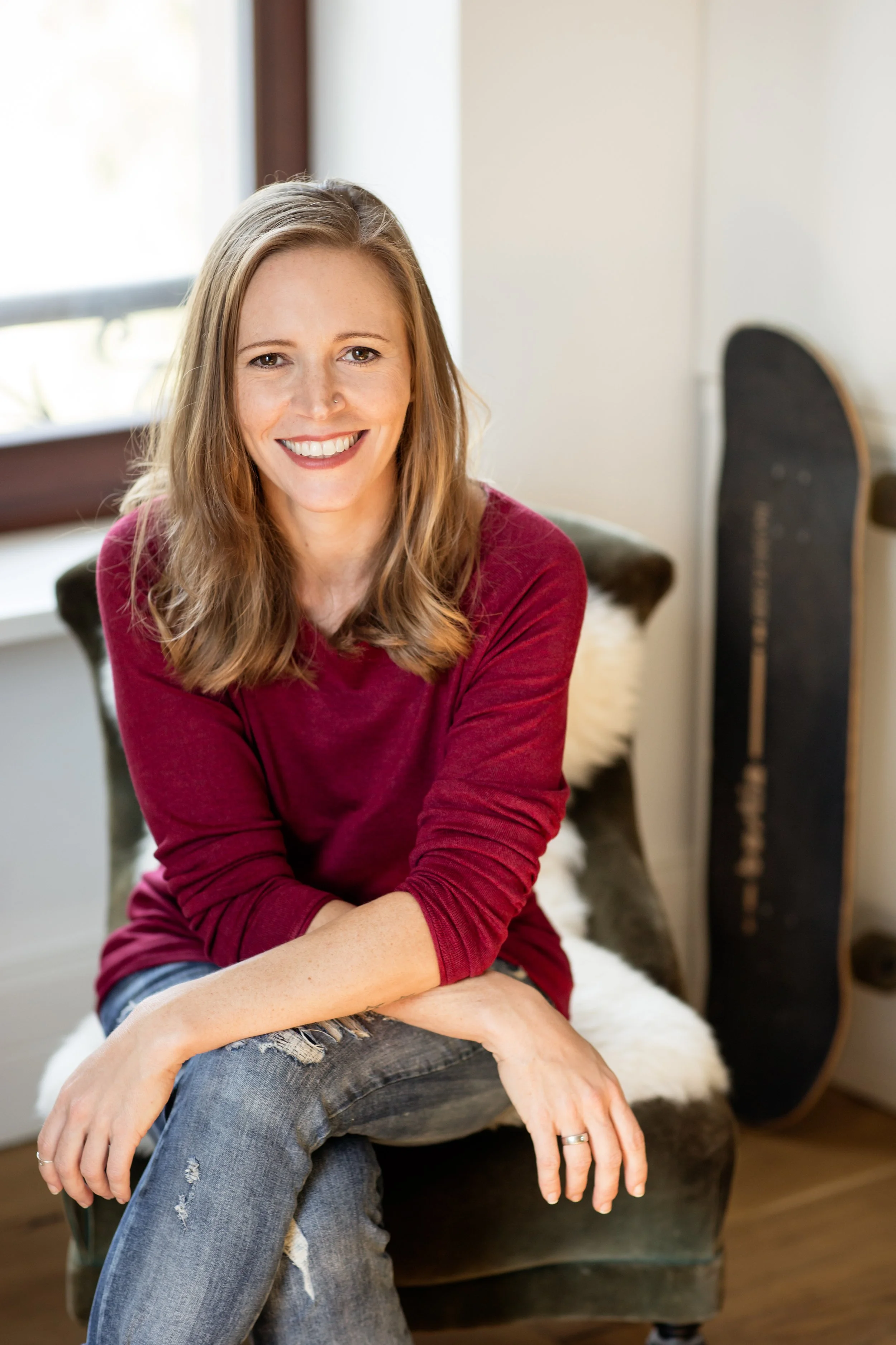 A woman with shoulder-length light brown hair, wearing a red long-sleeve shirt and ripped jeans, sitting on a vintage-style armchair with a sheepskin throw. She is smiling and sitting in a cozy, well-lit room near a window with natural light.