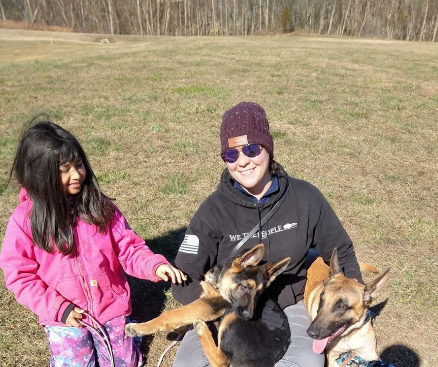 A woman with purple sunglasses and a purple beanie smiling while kneeling in a grassy field, holding two dogs, with a young girl in a pink jacket reaching out to pet one of the dogs.