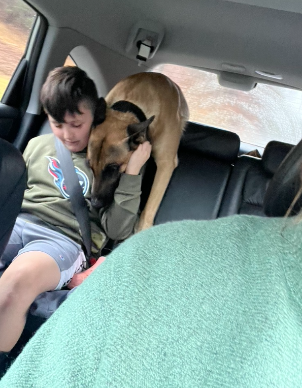 A boy in a beige jacket sitting in the backseat of a car, hugging and kissing a large tan and black dog that is standing on the seat beside him. The dog appears to be comforting the boy.