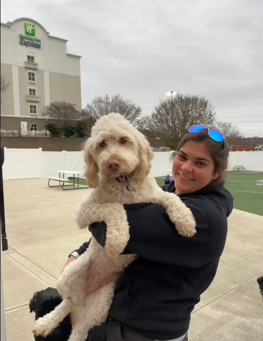 A young woman is smiling and holding a large, fluffy light-colored dog outside on a paved area near a sports field. The woman is wearing sunglasses on her head and a dark jacket. In the background, there is a white fence, trees, a hotel building, and