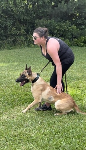A woman in black athletic wear crouches down to hold a Belgian Malinois dog on a leash, on a grassy field with trees in the background.