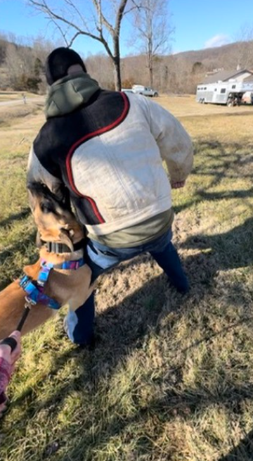 A person in a gray jacket with black, red, and white accents is walking or running outdoors on a grassy area. A dog on a leash is in the foreground, wearing a colorful collar.