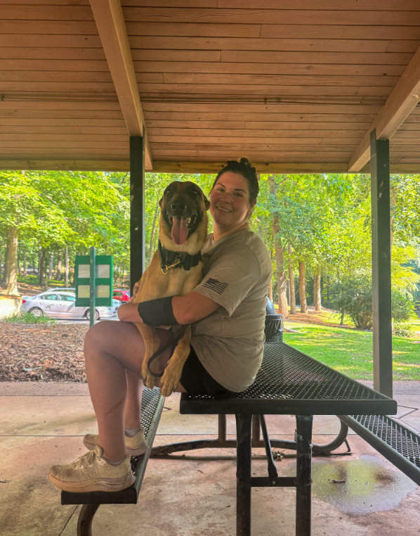 A woman sitting on a black metal picnic table holding a large happy dog in her lap under a pavilion in a park with green trees in the background.