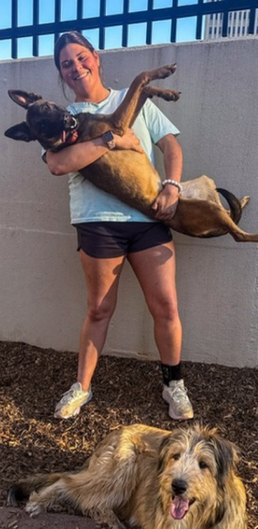 A woman holding a large dog in her arms, both smiling, with another dog lying on the ground nearby, in an outdoor setting with a concrete wall and metal fence in the background.