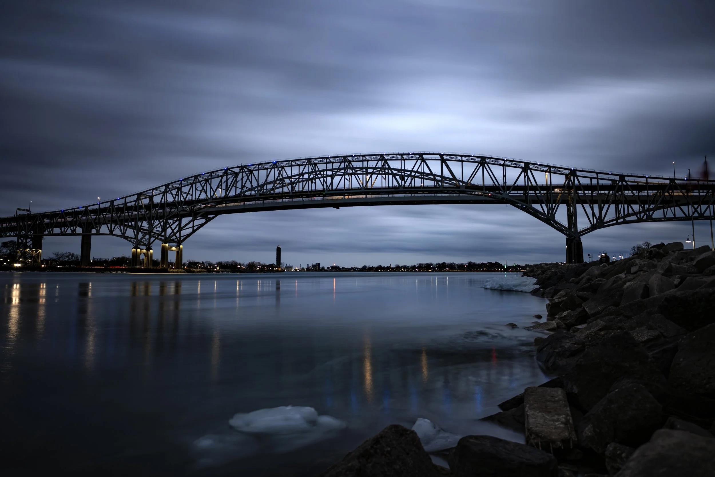 Long-exposure blue hour photo of the Bluewater Bridge over the St. Clair River during night, with clouds moving across the sky and city lights reflecting on the water's smooth surface.