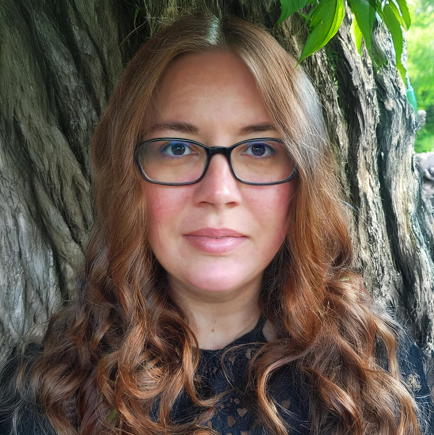 A woman with long, curly red hair and glasses standing in front of a large tree trunk and green foliage.