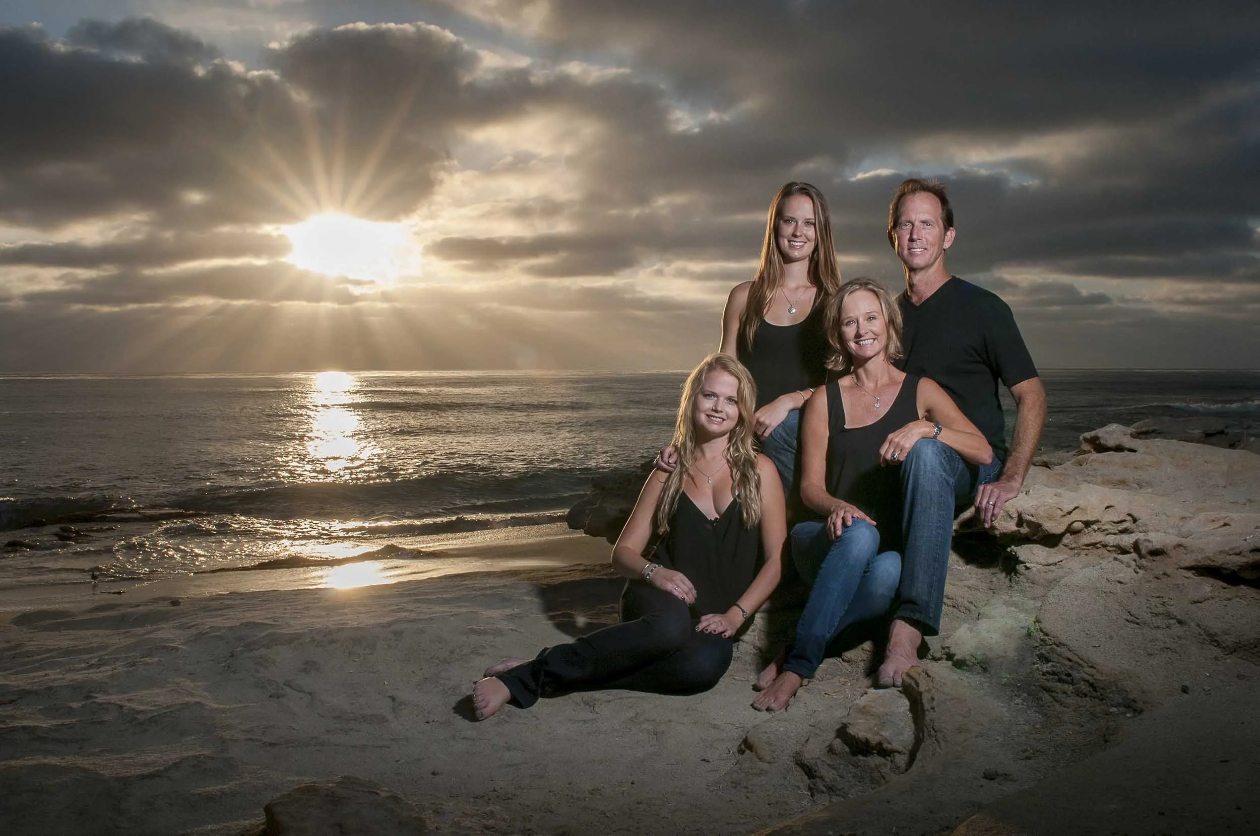 A family of four enjoying a sunset on the beach. They are sitting on rocks with the ocean in the background, under a partly cloudy sky with sunlight breaking through.