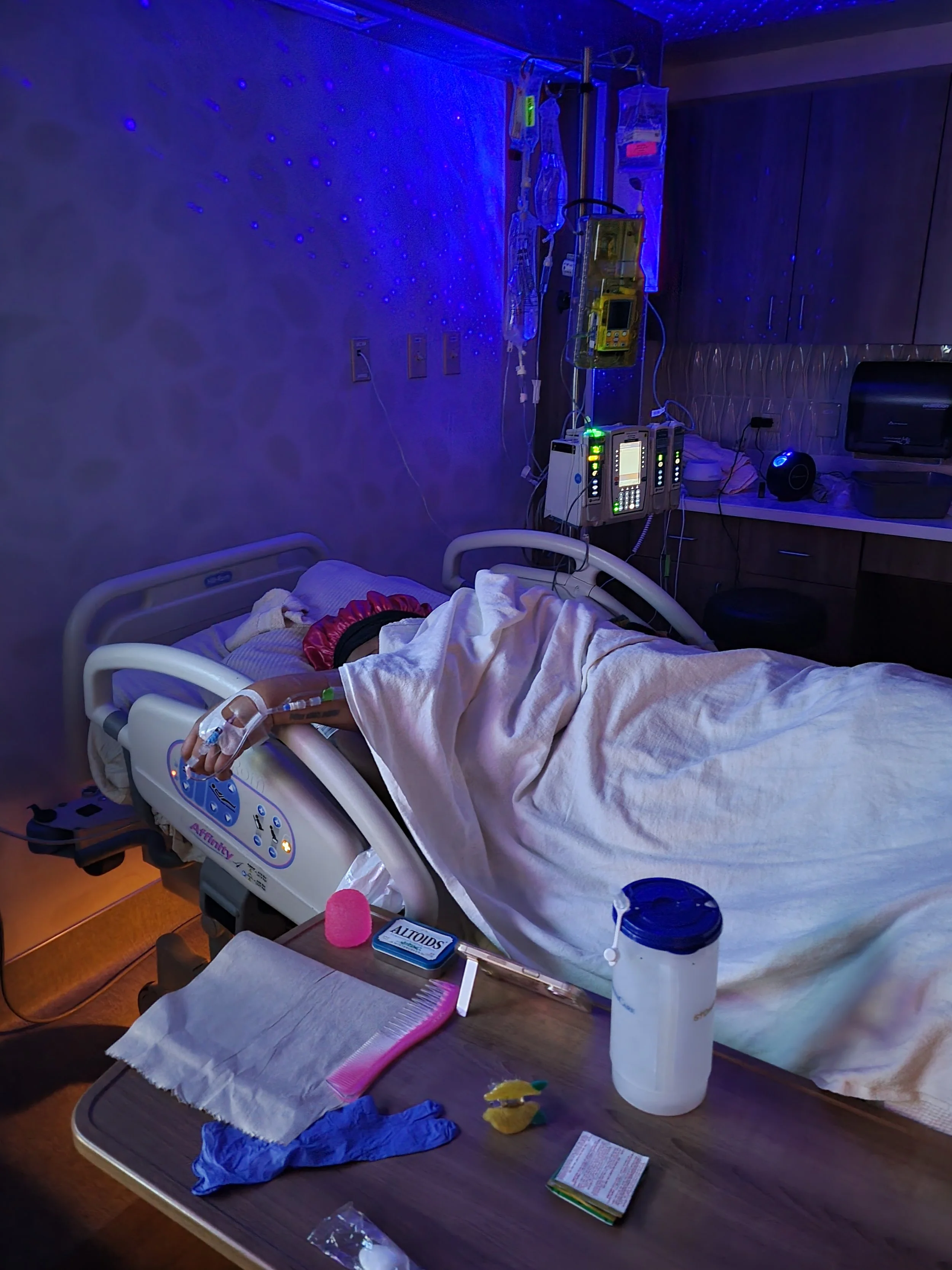 Hospital room with patient lying in bed connected to medical devices, IV drip, and monitors. Nighttime with blue and purple lighting, medical supplies on a tray, and a closed cabinet.