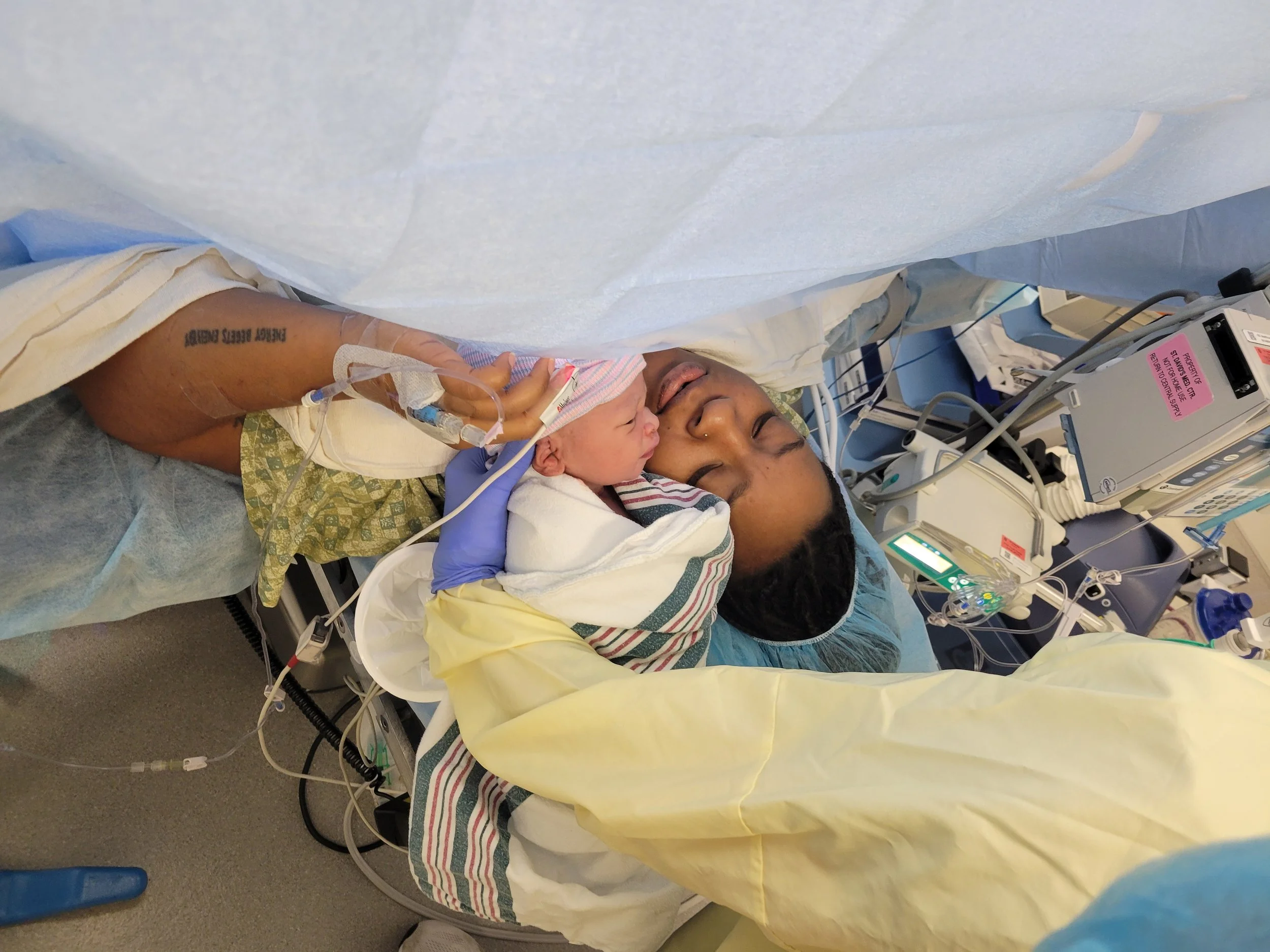 A woman lying in a hospital bed with a newborn baby on her chest, surrounded by medical equipment and monitoring devices.