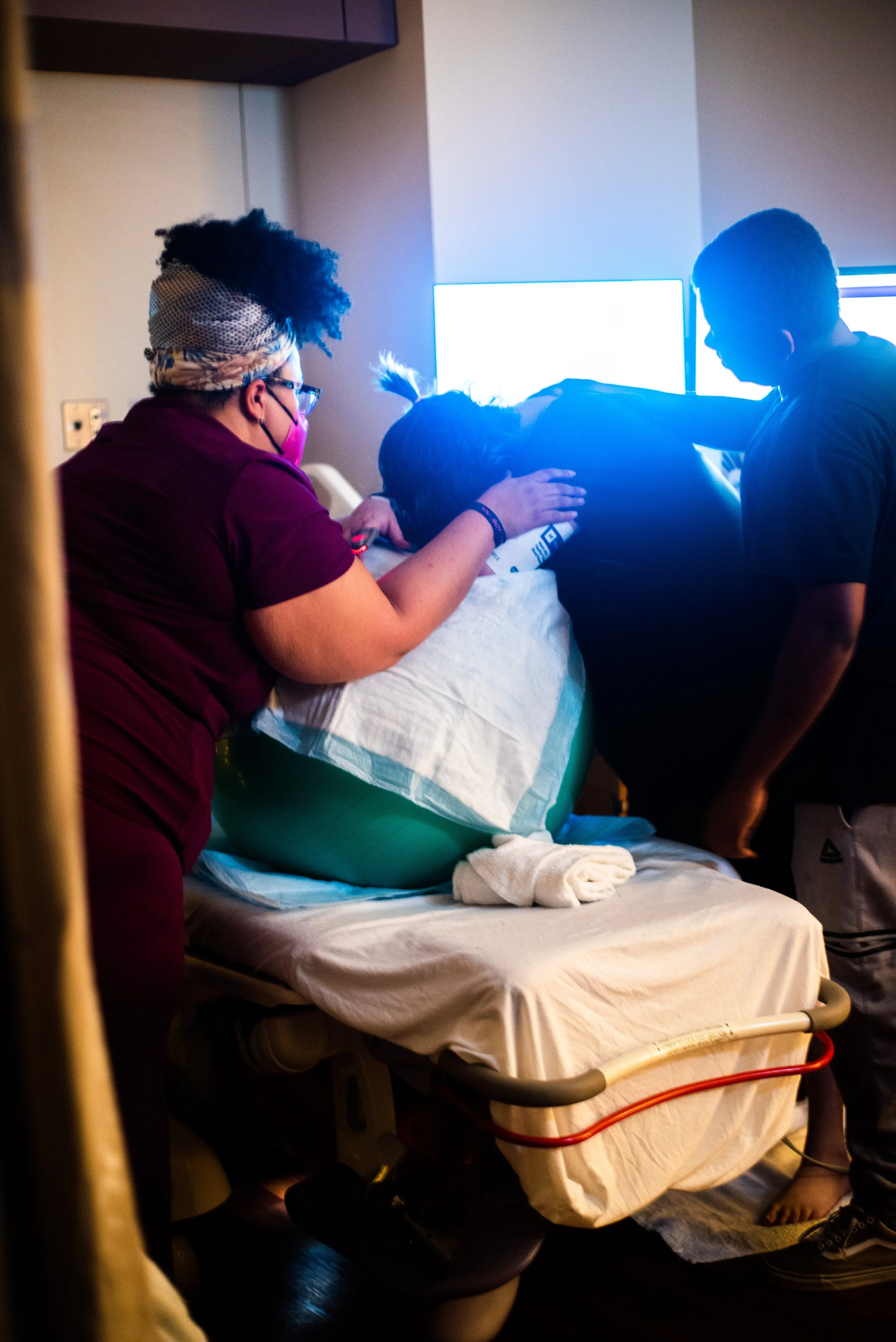 A healthcare team attends to a patient lying in a hospital bed, with two monitors in the background and a healthcare worker wearing a face mask and glasses beside the bed.