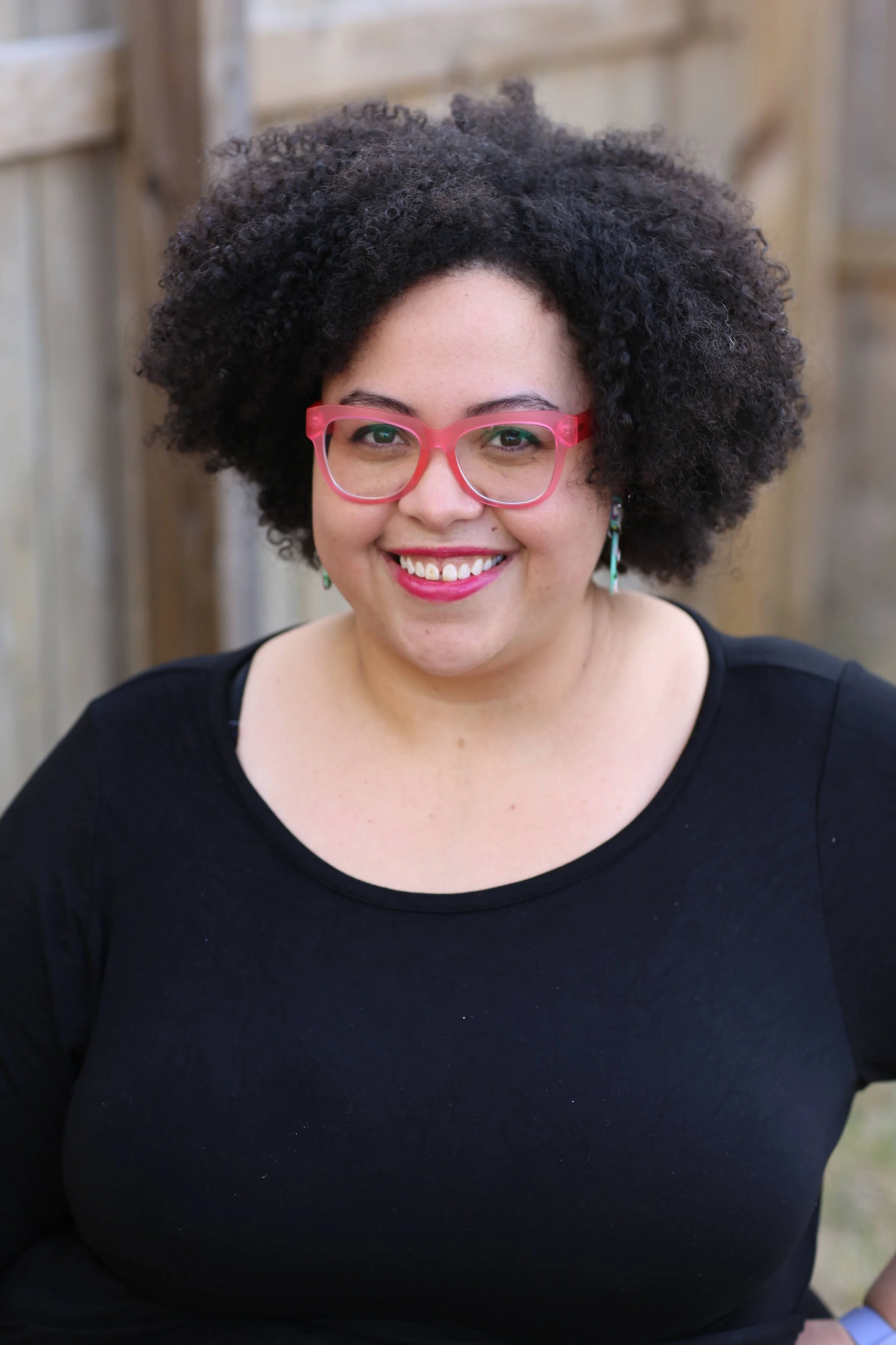 A woman with curly afro hair, pink glasses, and earrings smiling outdoors with a wooden fence in the background.