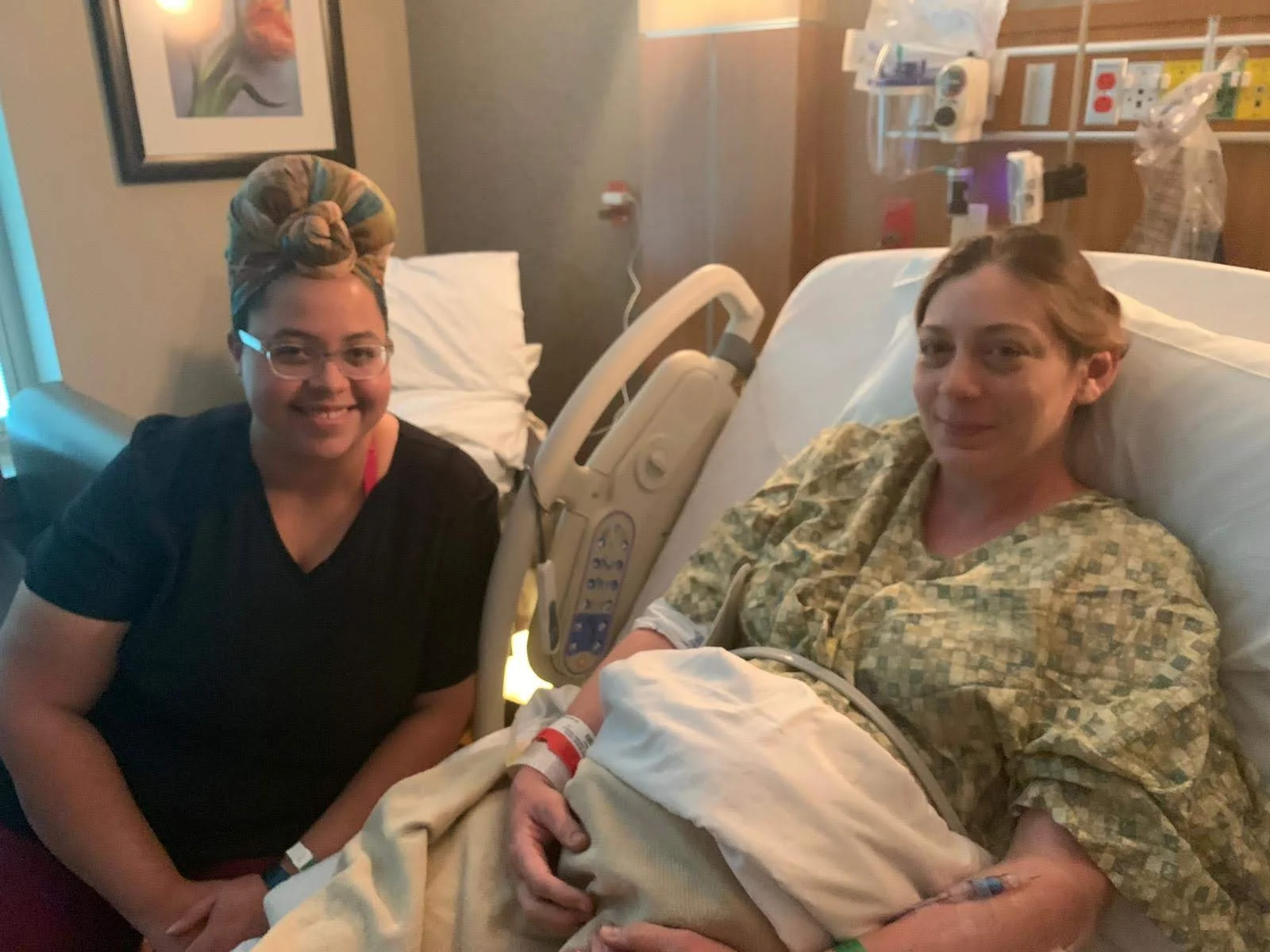 A woman in a hospital bed with a yellow-green patterned gown, holding her newborn baby, Asian woman with short brown hair, sitting beside her in a hospital room. A woman with glasses and a colorful headscarf kneels beside them, smiling.