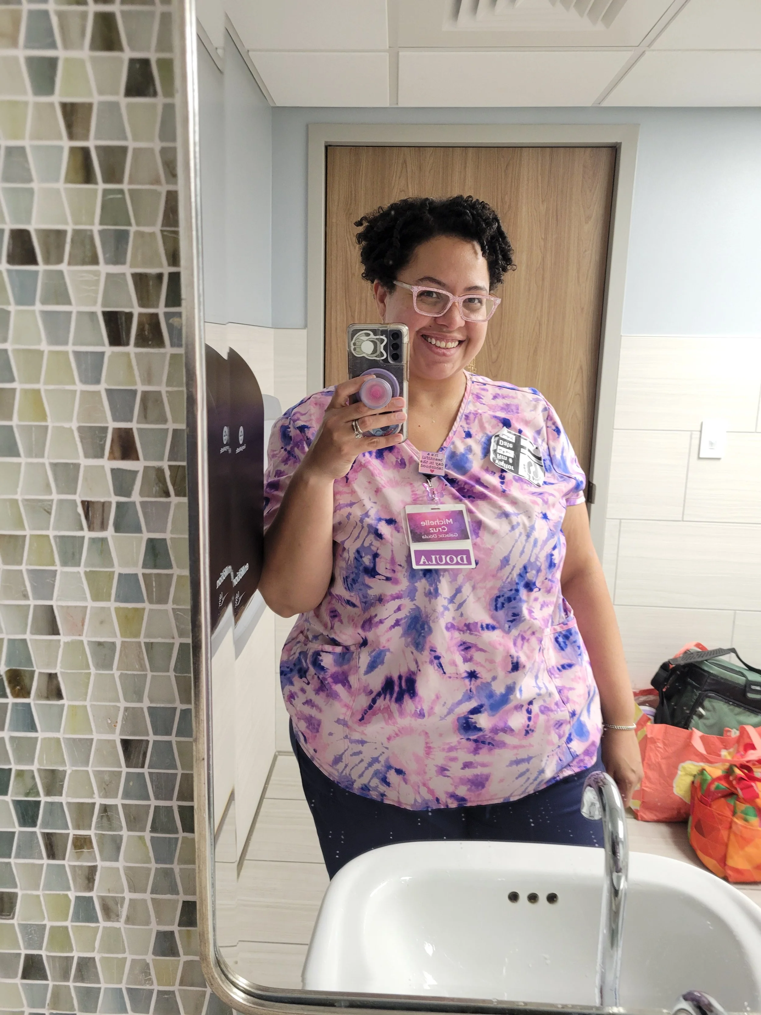 Smiling woman wearing pink glasses and a tie-dye scrub top taking a selfie in a bathroom mirror.