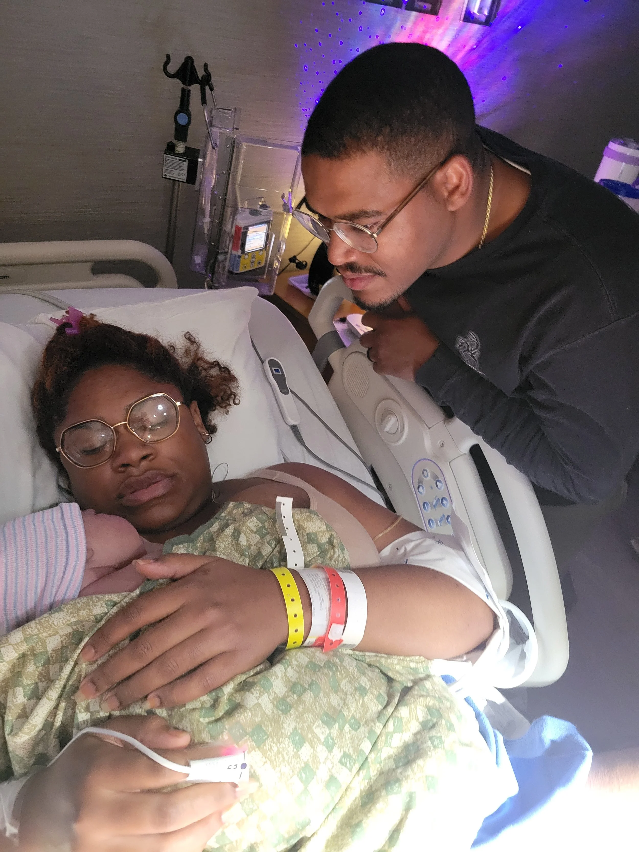 A woman lying in a hospital bed with a hospital gown, wearing glasses, and medical wristbands. A man with glasses is leaning over her, holding her hand, in a hospital room with medical equipment and purple lighting in the background.