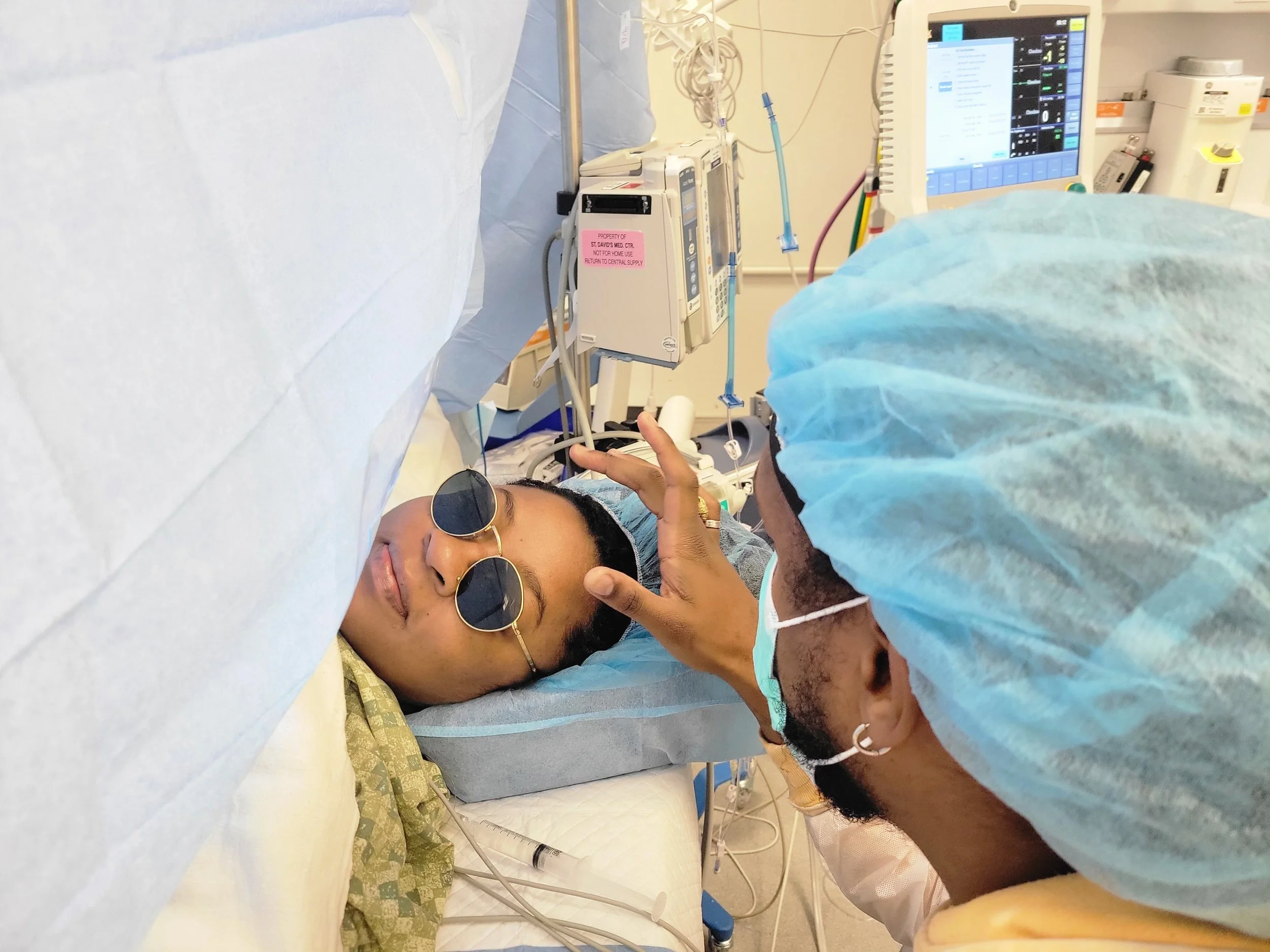 A person lying in a hospital bed wearing glasses and a patterned hospital gown, with a medical professional attending to them, wearing a blue surgical cap and gown, in a hospital room with medical monitors and equipment.