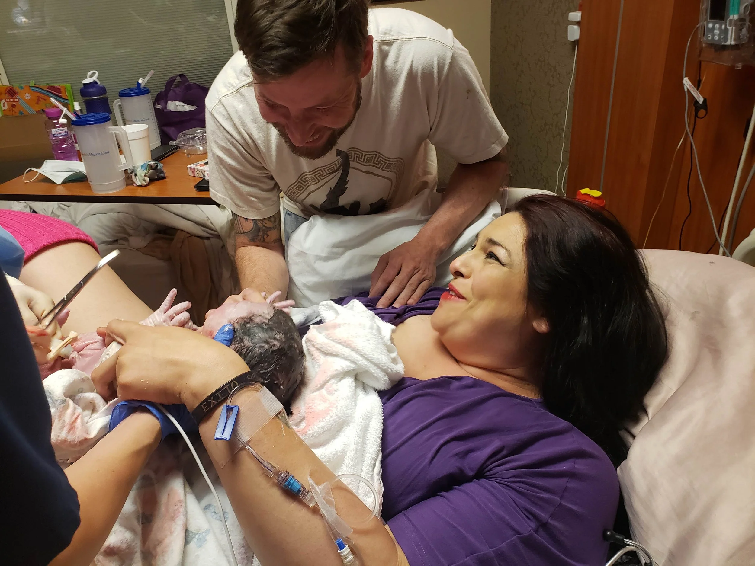 A woman lying in a hospital bed, smiling, with a man leaning over her, holding her hand during childbirth. Medical equipment and supplies are visible in the background.