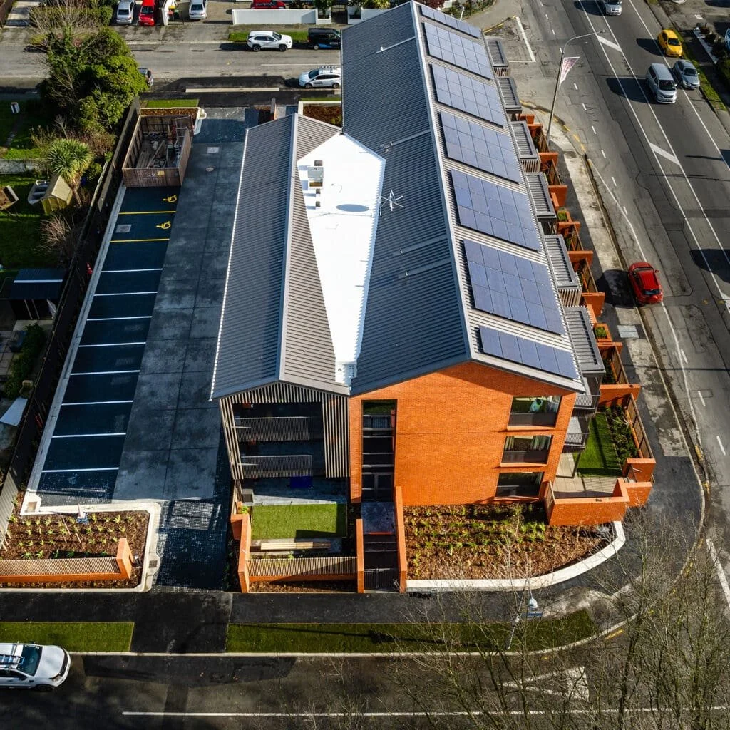 An aerial view of a modern multi-story building with solar panels on the roof, located at the corner of a street with parked cars and adjacent buildings.