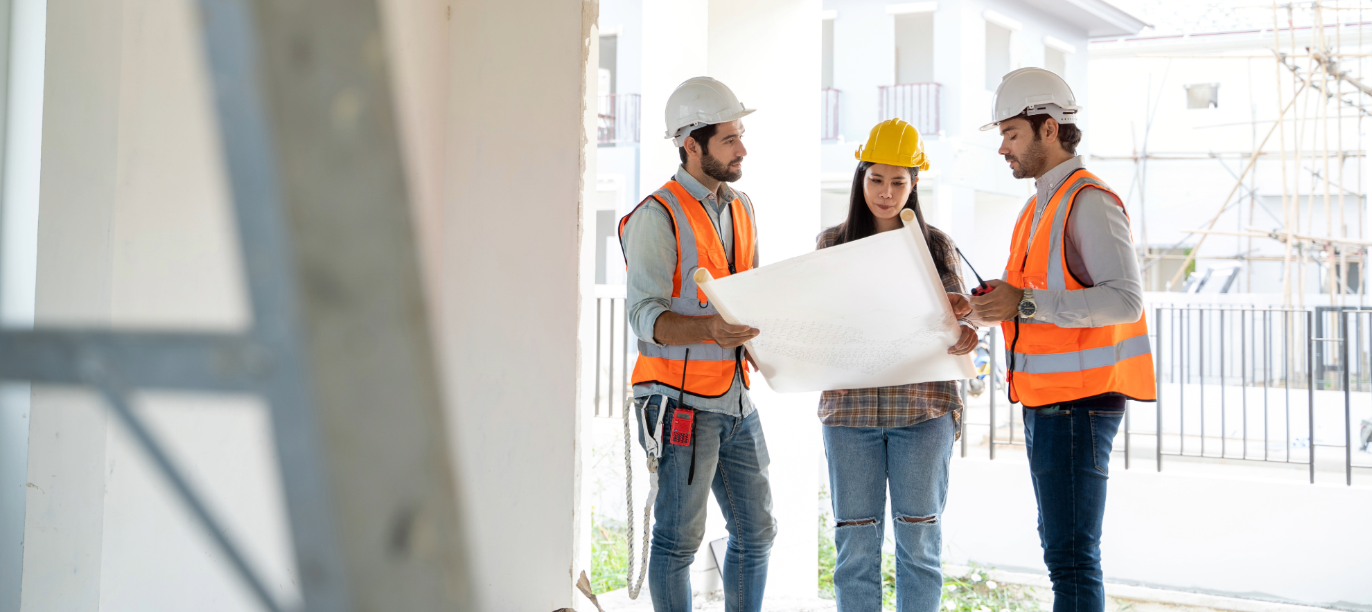 Three construction workers wearing safety helmets and orange safety vests reviewing blueprints on a construction site.