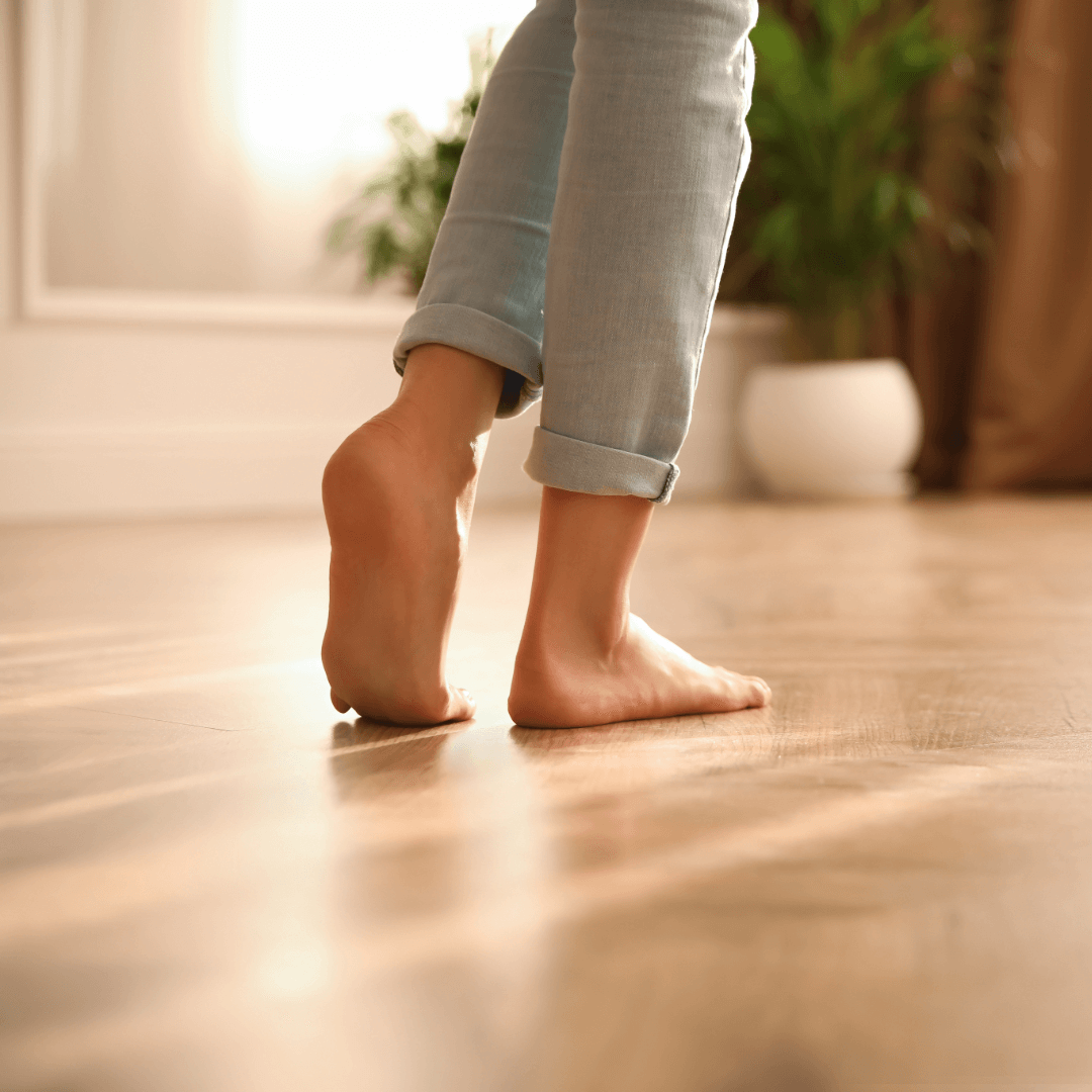 Person standing on toes barefoot on wooden floor, wearing rolled-up light-colored pants, with a blurred background including a potted plant.