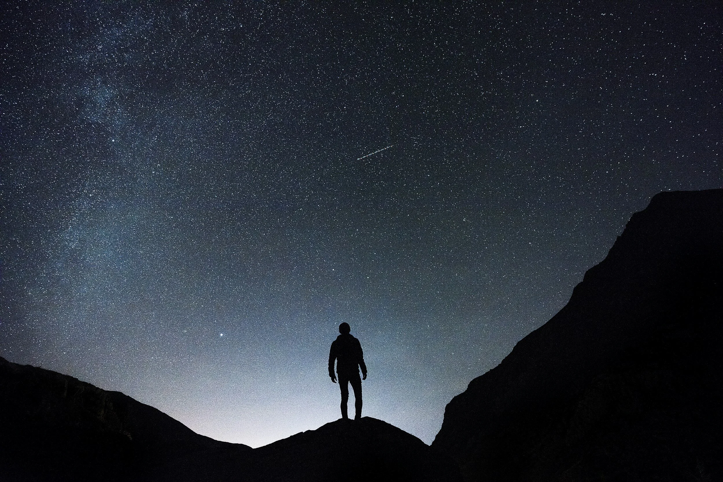 A person stands on a hill under a starry night sky with numerous stars and a visible shooting star or comet.