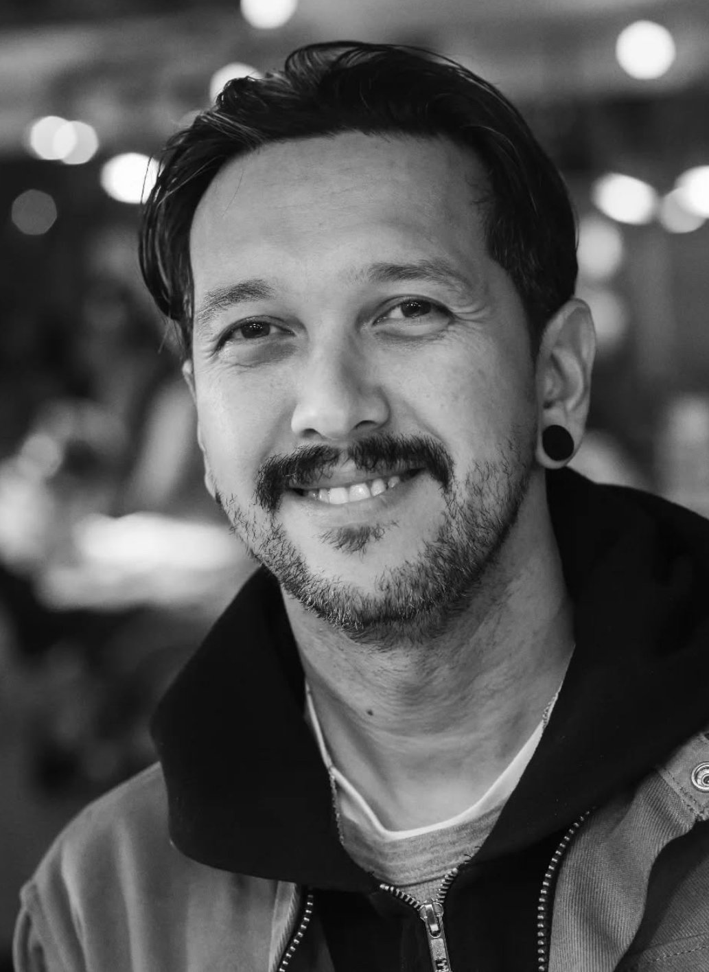 Close-up black and white portrait of a smiling man with short dark hair, goatee, and a mustache, wearing a jacket with a zipper and earrings, in an indoor setting with blurred background lights.
