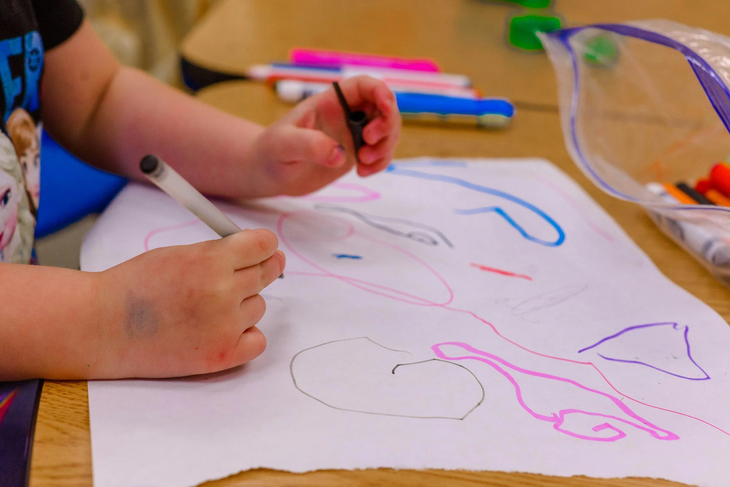 A child's hands drawing on white paper with colorful markers, with a small zippered plastic bag and various markers on a wooden table.